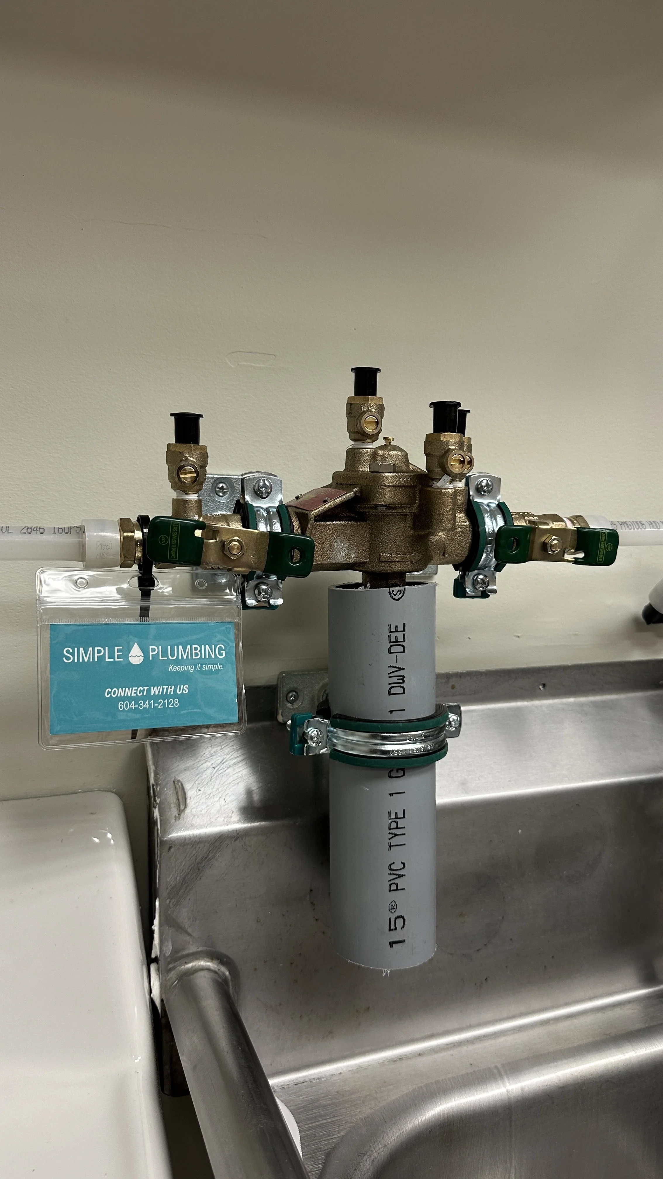Close-up of a plumbing valve system with PVC pipes attached, mounted on a stainless steel sink and wall, including a blue branding tag from Simple Plumbing.