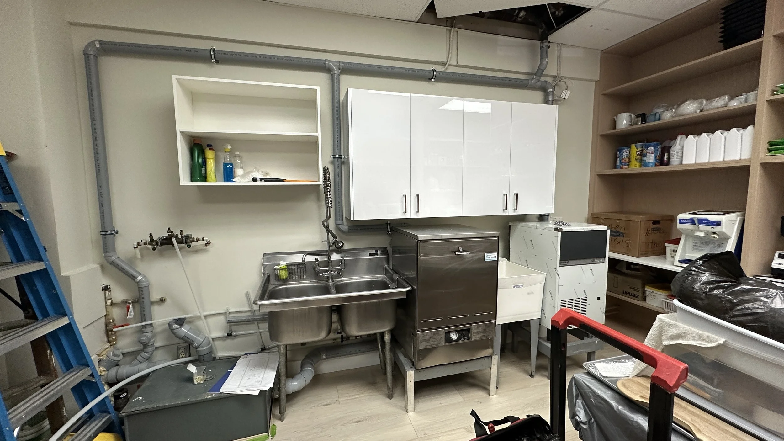 A utility or cleaning room with a stainless steel double sink, white cabinets, shelves with cleaning supplies, a box, a black trash bag, and various tools and equipment.