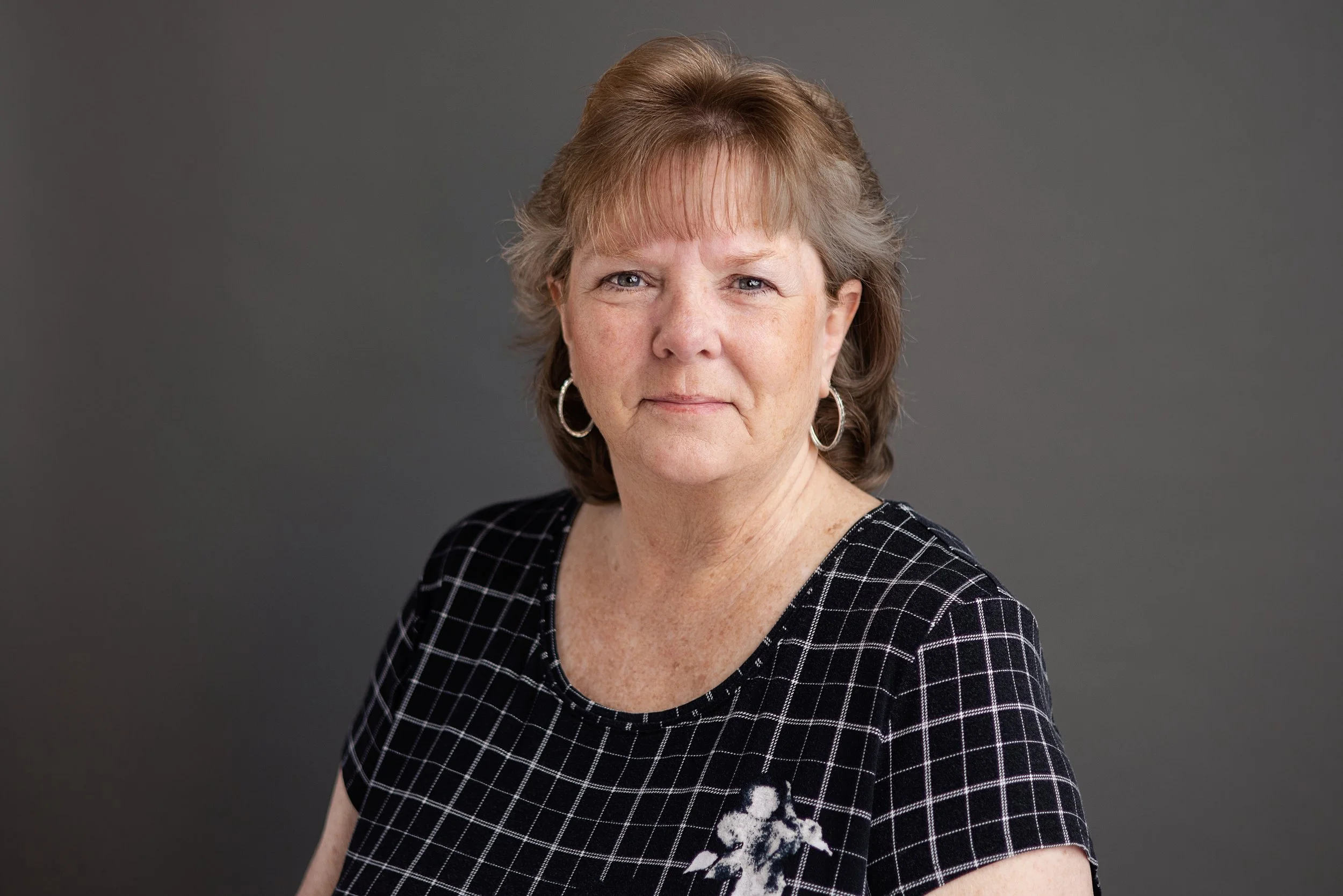 A middle-aged woman with short brown hair and blue eyes wearing a black dress with a white checkered pattern, silver hoop earrings, and a small floral design on her dress, posed against a plain gray background.