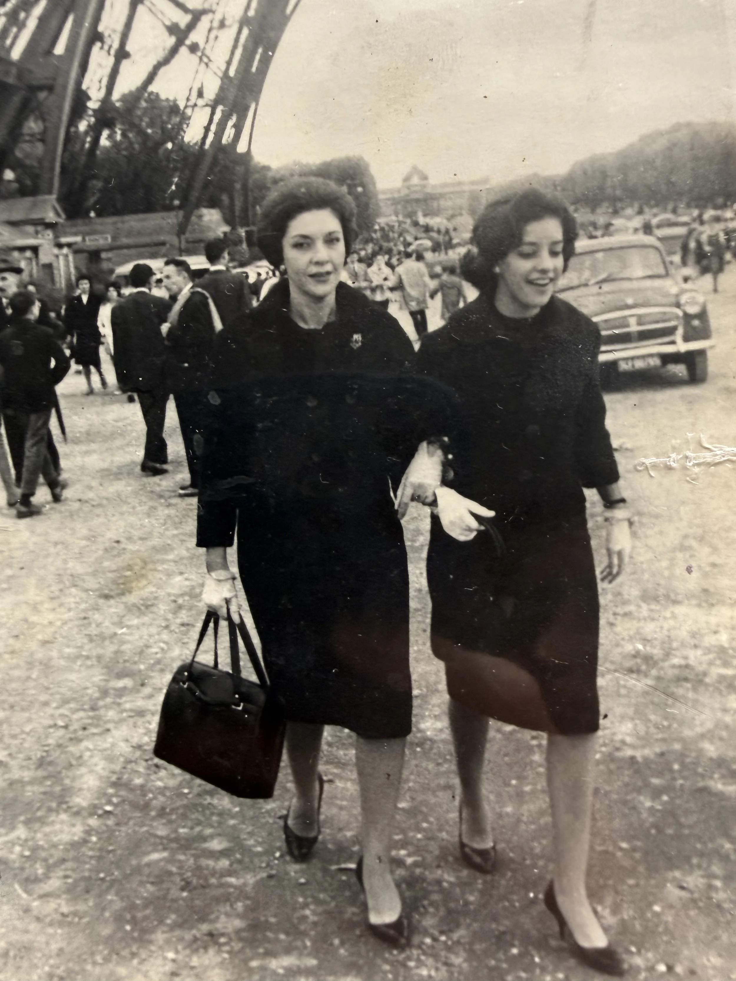 Dos mujeres vestidas con ropa oscura caminando con caballos en una feria o parque, con gente y carros en el fondo, imagen en blanco y negro.