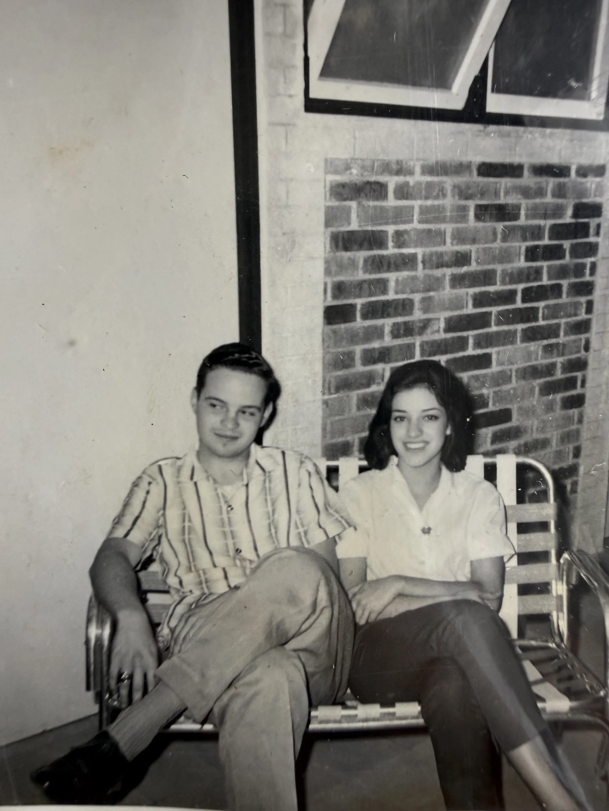 Dos personas, un hombre y una mujer, sentados juntos en un banco de madera en un patio con pared de ladrillos y paredes blancas, en una foto en blanco y negro.