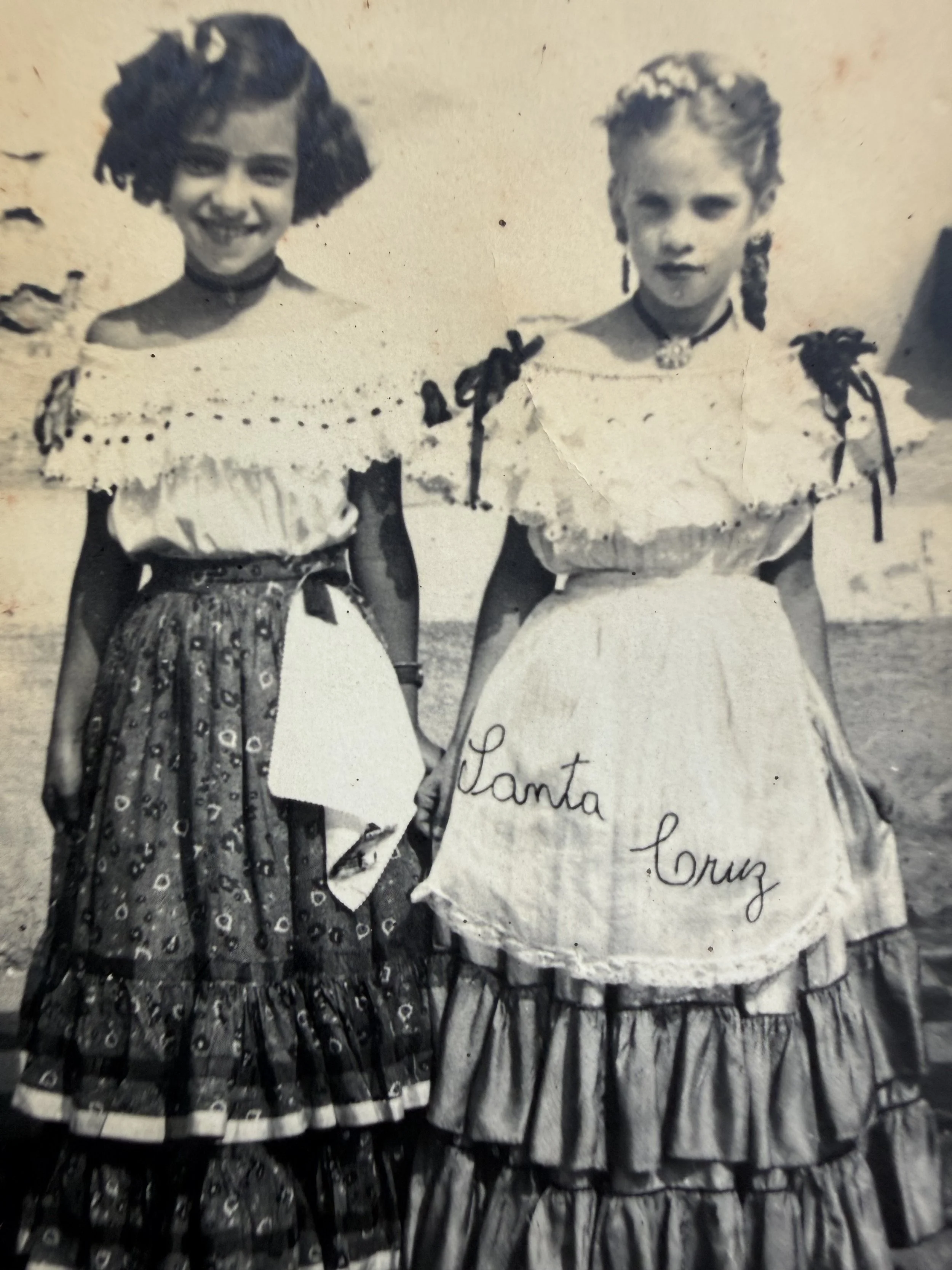 Dos mujeres jóvenes con trajes típicos de fiesta, sonriendo y sosteniendo una pancarta que dice 'Santa Cruz'.