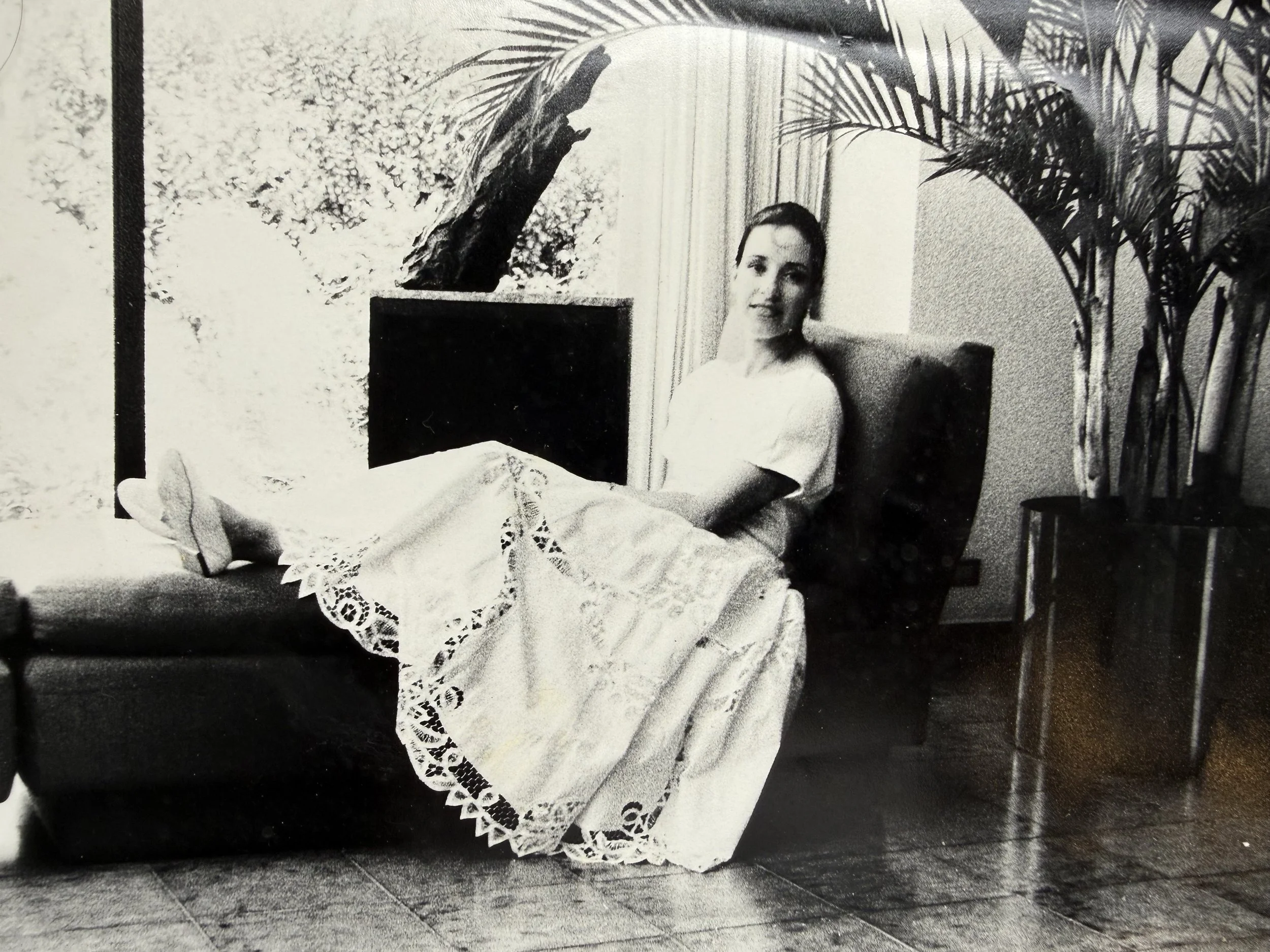 Retrato de una mujer sentada en una sala, vestida con un vestido largo de encaje, rodeada de plantas de interior