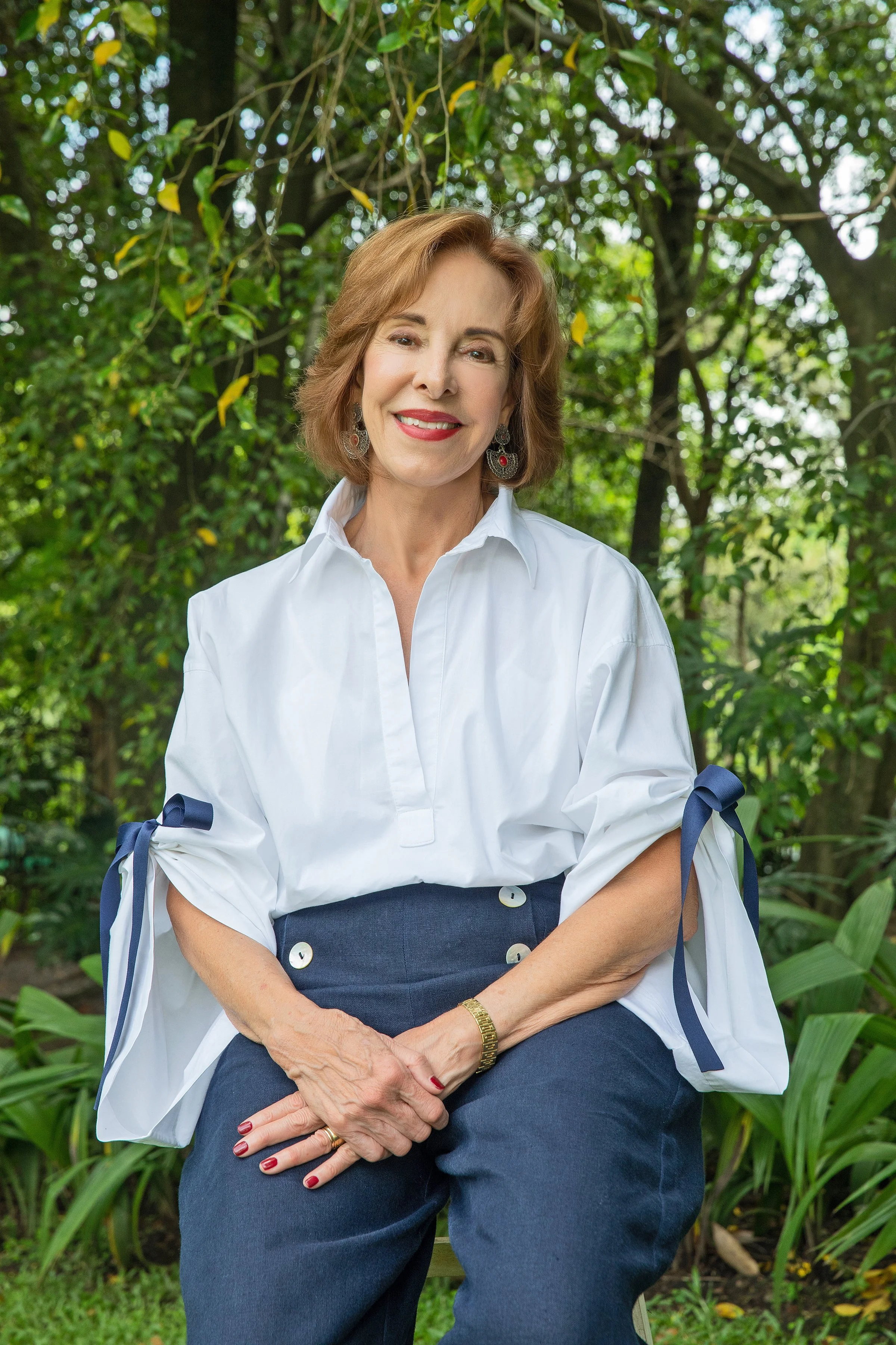Mujer de cabello castaño, con vestido oscuro y blusa blanca, posando con sonrisa en un entorno natural con árboles y plantas verdes.