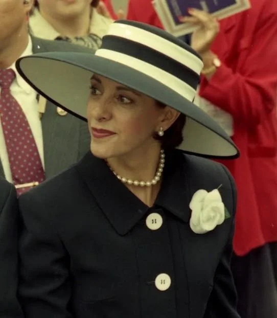Mujer vestida con abrigo negro, sombrero amplio en colores beige y negro, con una flor blanca en el saco, y collar de perlas, en un evento social con varias personas en el fondo.