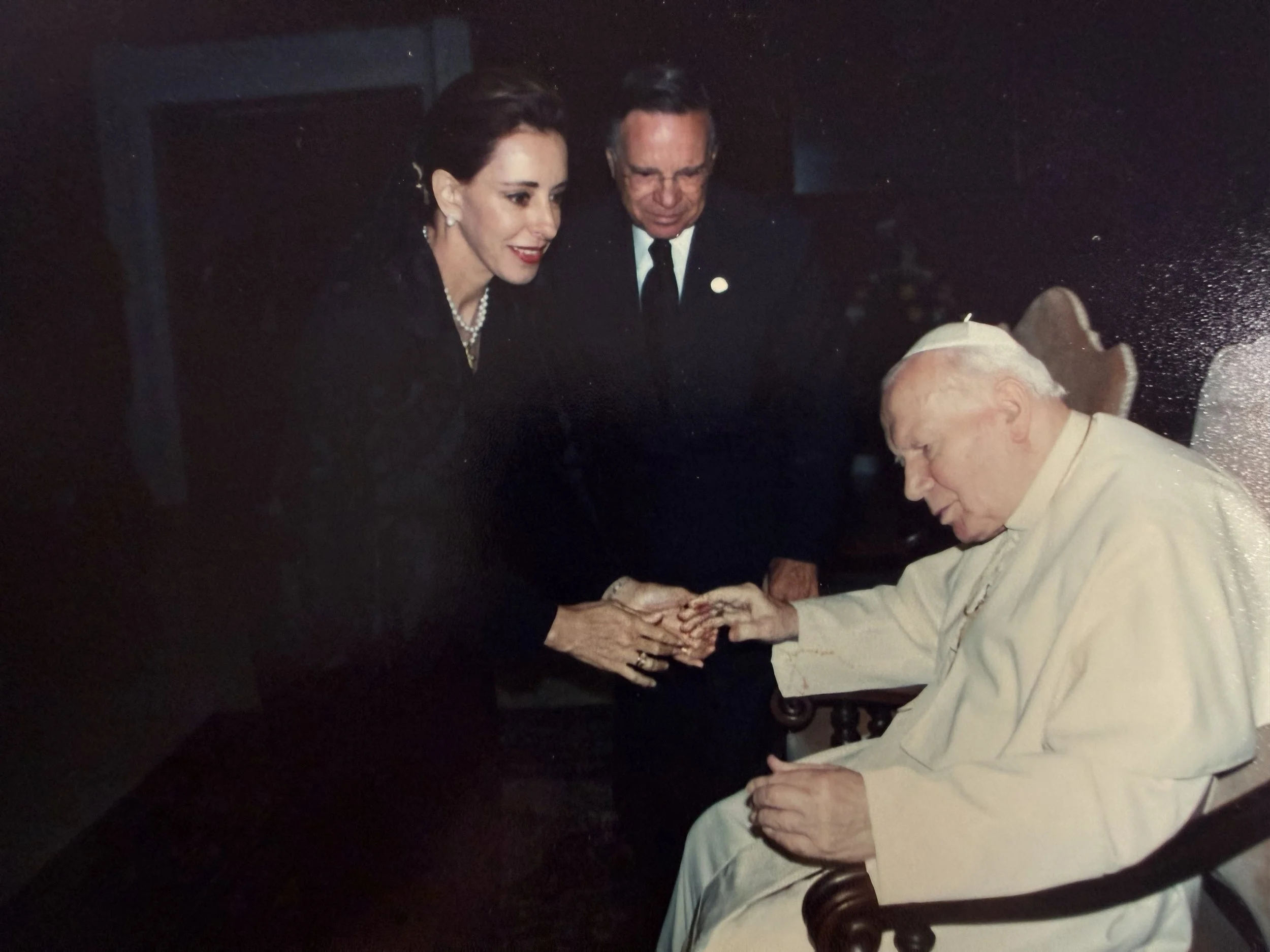 El Papa Francisco con dos personas mayores, una mujer y un hombre, en una reunión informal. La mujer tiene cabello oscuro, lleva pendientes y un collar de perlas, está sonriendo y usa ropa oscura. El hombre tiene cabello gris, usa gafas, un traje con