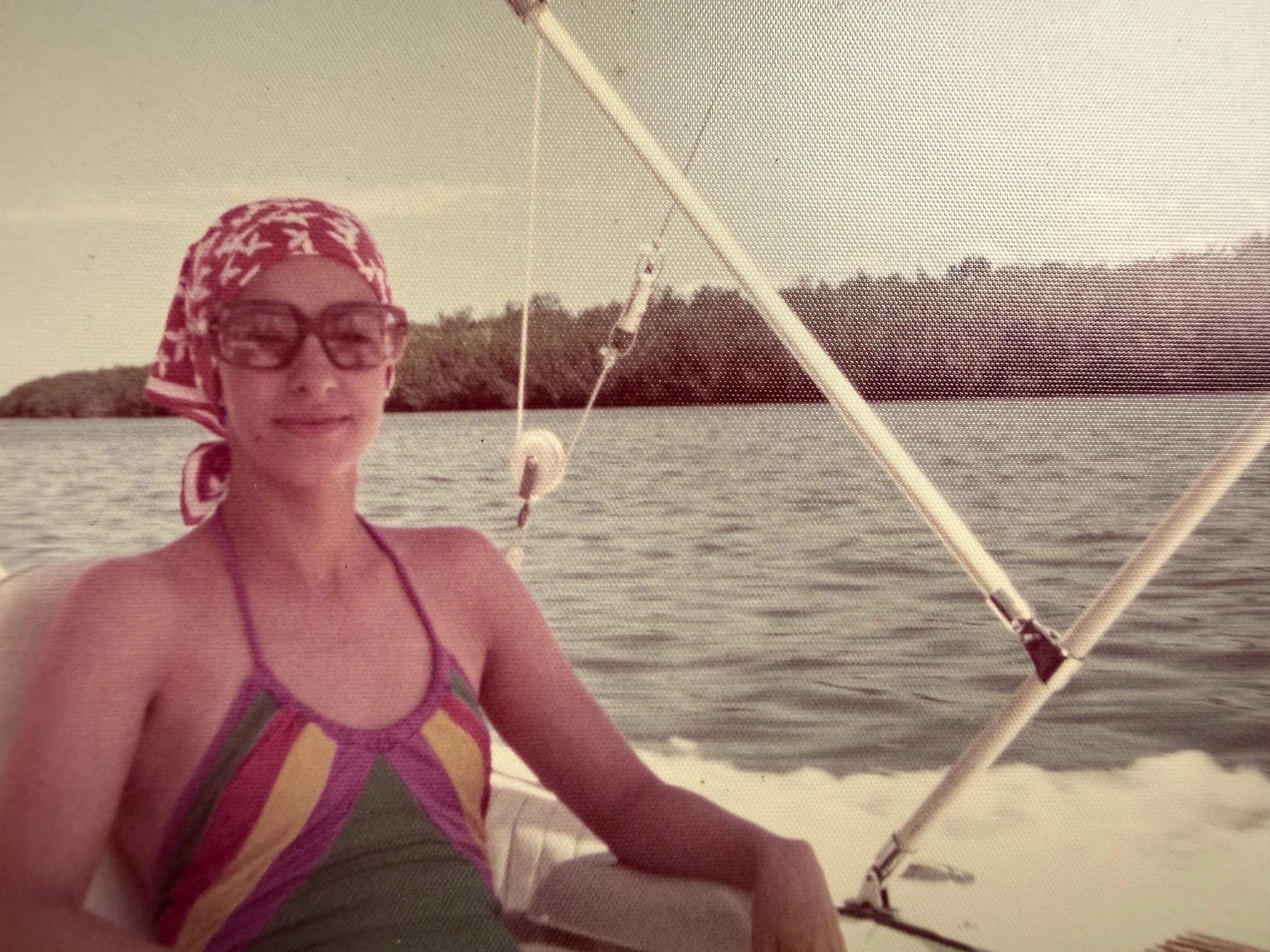 Una mujer de cabello rizado con pañuelo en la cabeza, gafas y bikini, pescando en un lago con vegetación en el fondo.