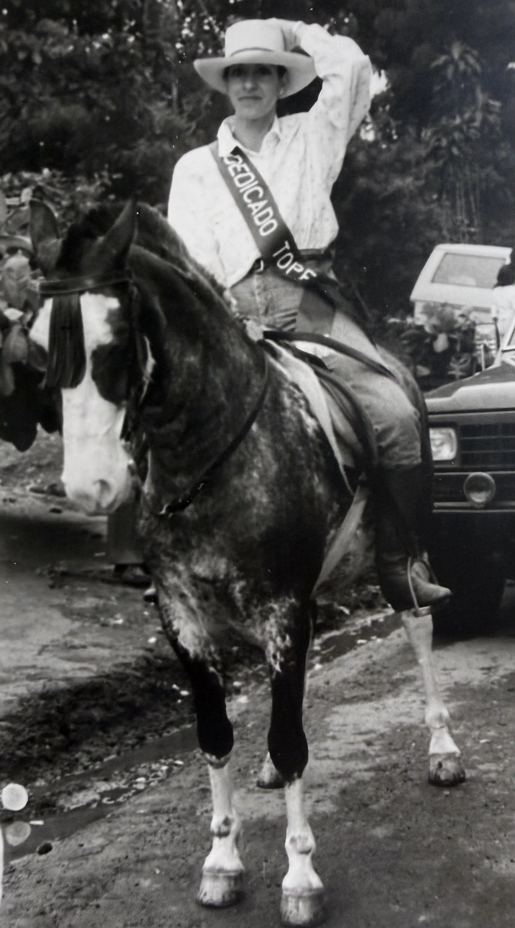 Una persona vestida con camisa y sombrero grande, montada en un caballo, usando una banda con la palabra 'GEOGRAFIA' y en el fondo hay autos y árboles.