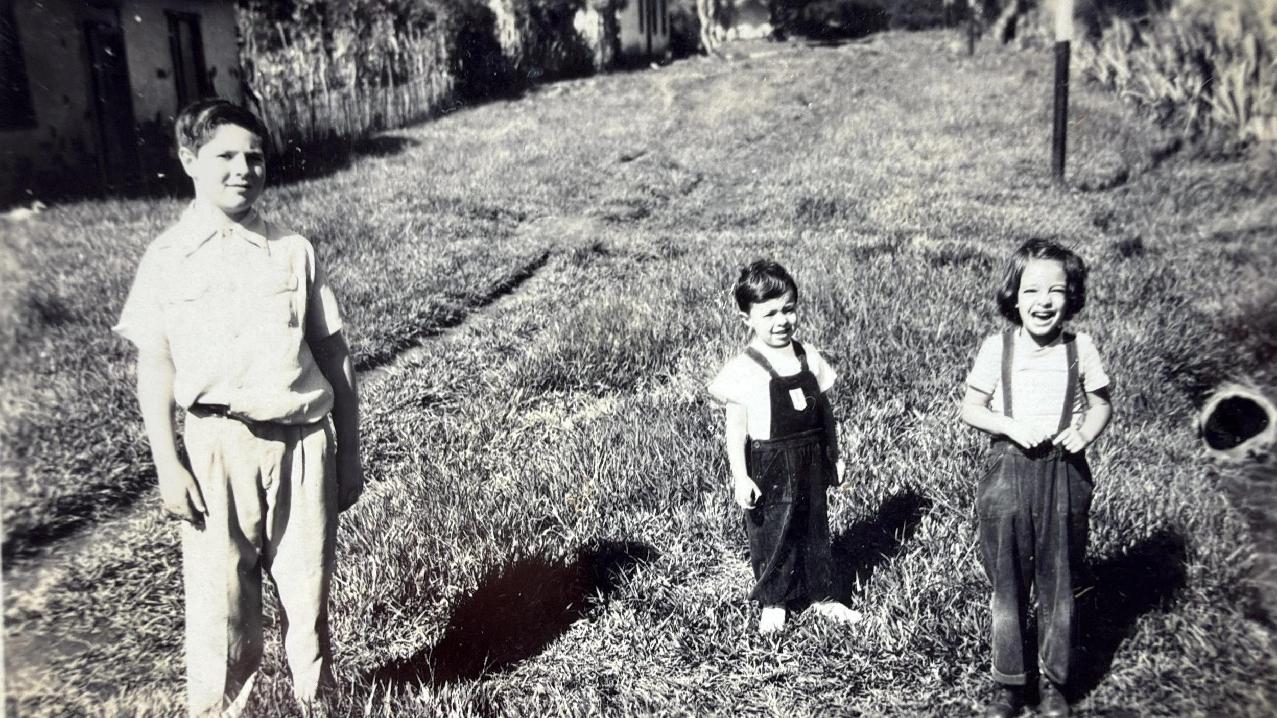 Una fotografía en blanco y negro de tres niños y una niña de pie en un campo de césped. La niña a la izquierda lleva camisa de manga larga y pantalones claros. El niño en el centro lleva una camiseta blanca y overoles oscuros. La niña a la derecha ll
