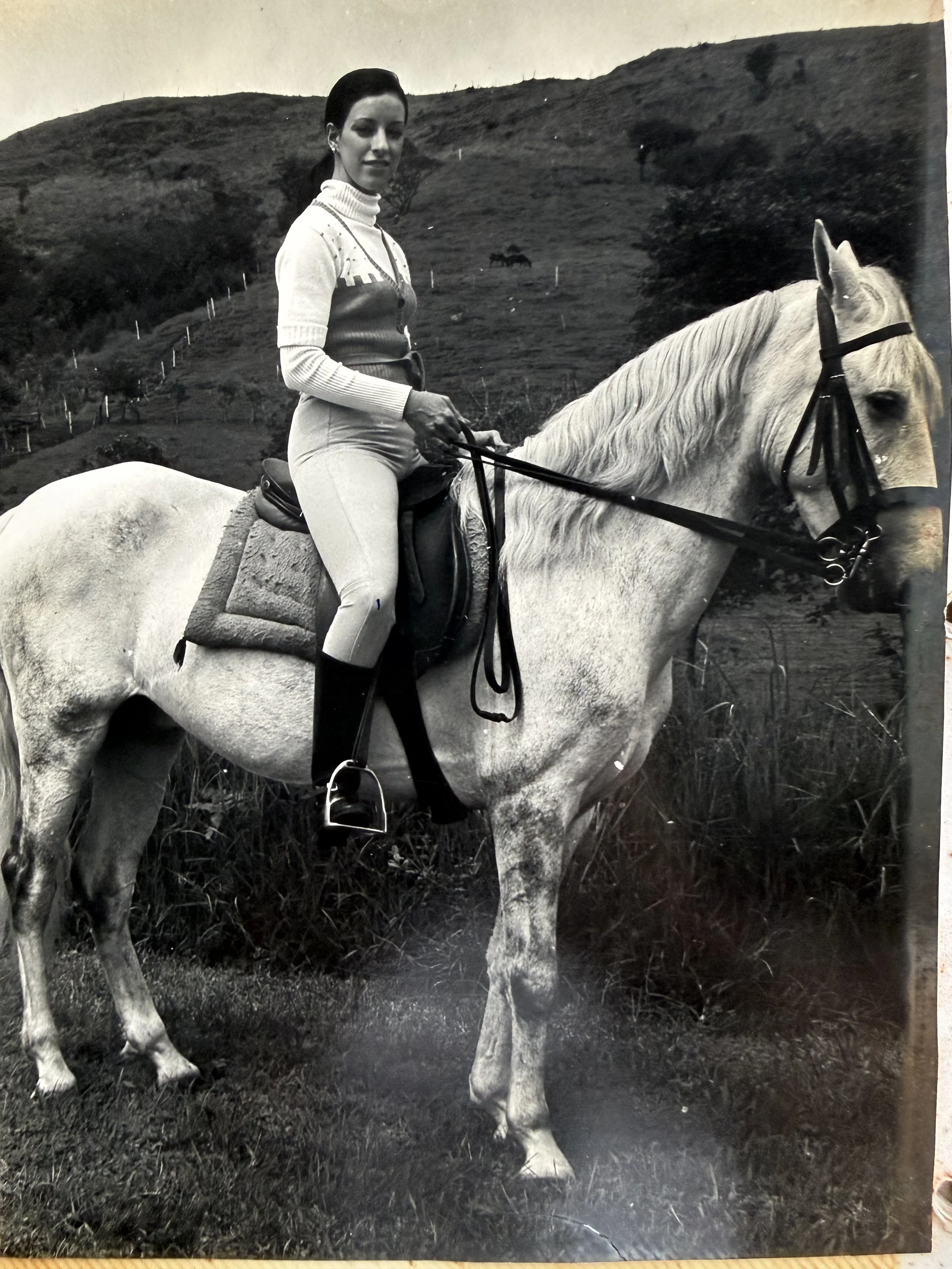 Mujer montando un caballo en un paisaje montañoso con césped y árboles, en una fotografía en blanco y negro.