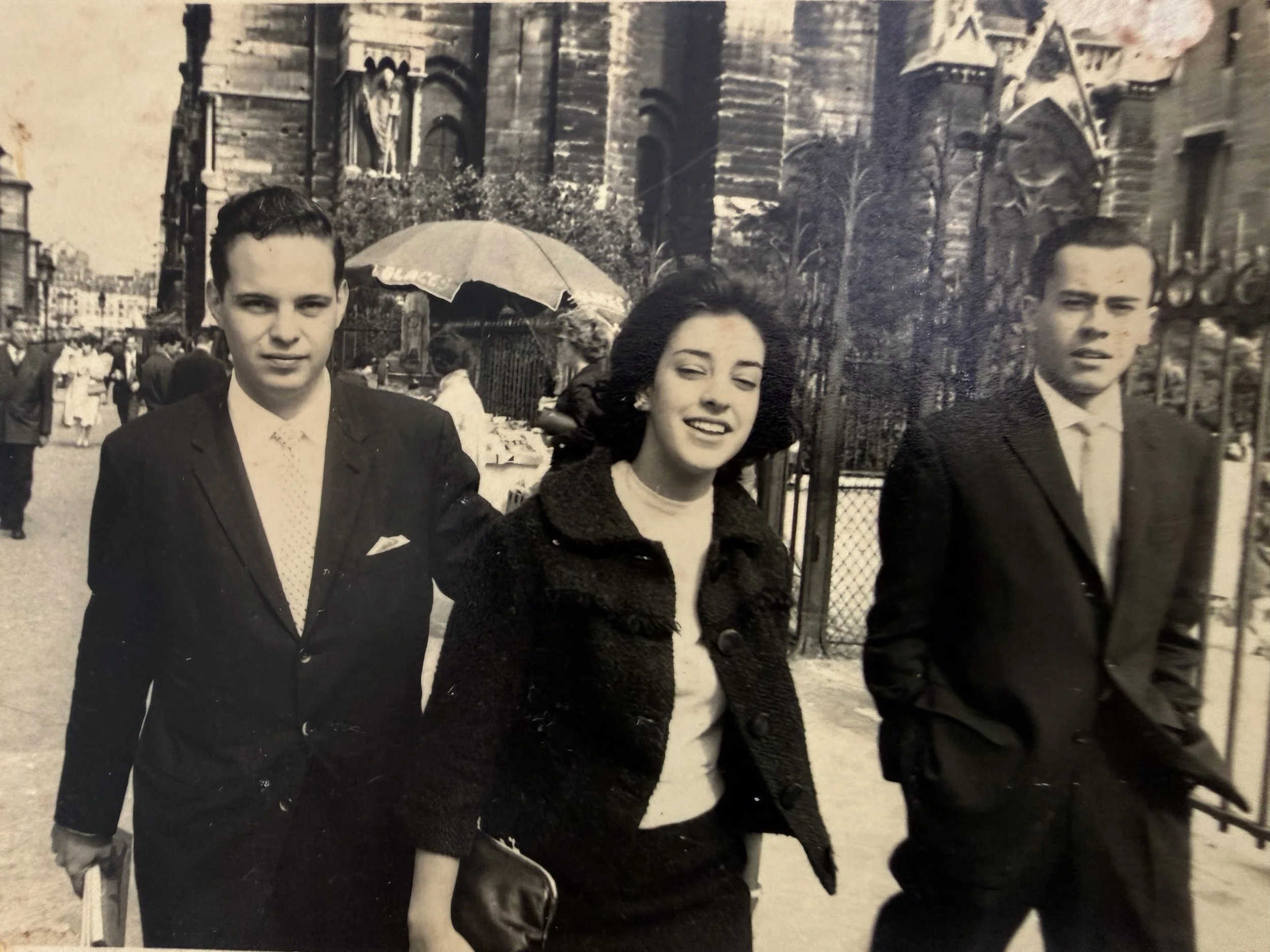 Fotografía en blanco y negro de un grupo de personas, en el centro dos hombres con trajes y una mujer con vestido, en un entorno urbano posiblemente en una ciudad europea antigua, con edificaciones históricas y un parque o calle con reja de fondo.