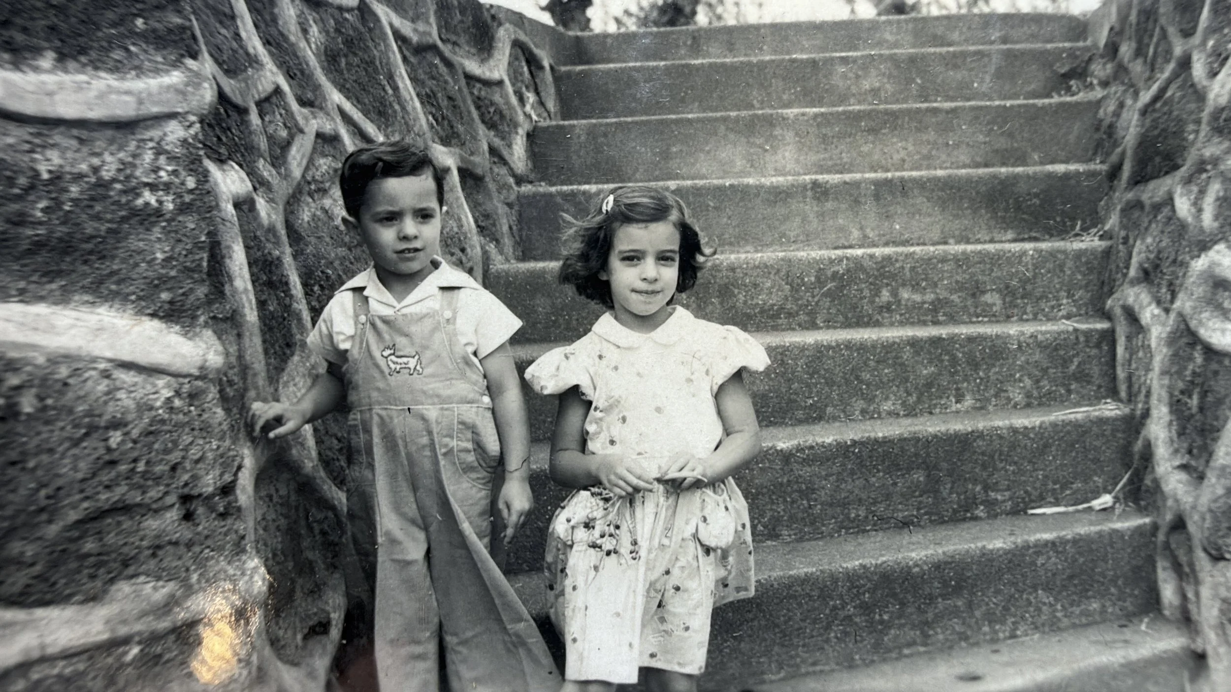 Dos niños pequeños, un niño y una niña, posando en las escaleras de piedra, la niña lleva puesto un vestido de vestido de verano y el niño lleva puesto un overol con una camiseta debajo, en una fotografía en blanco y negro.