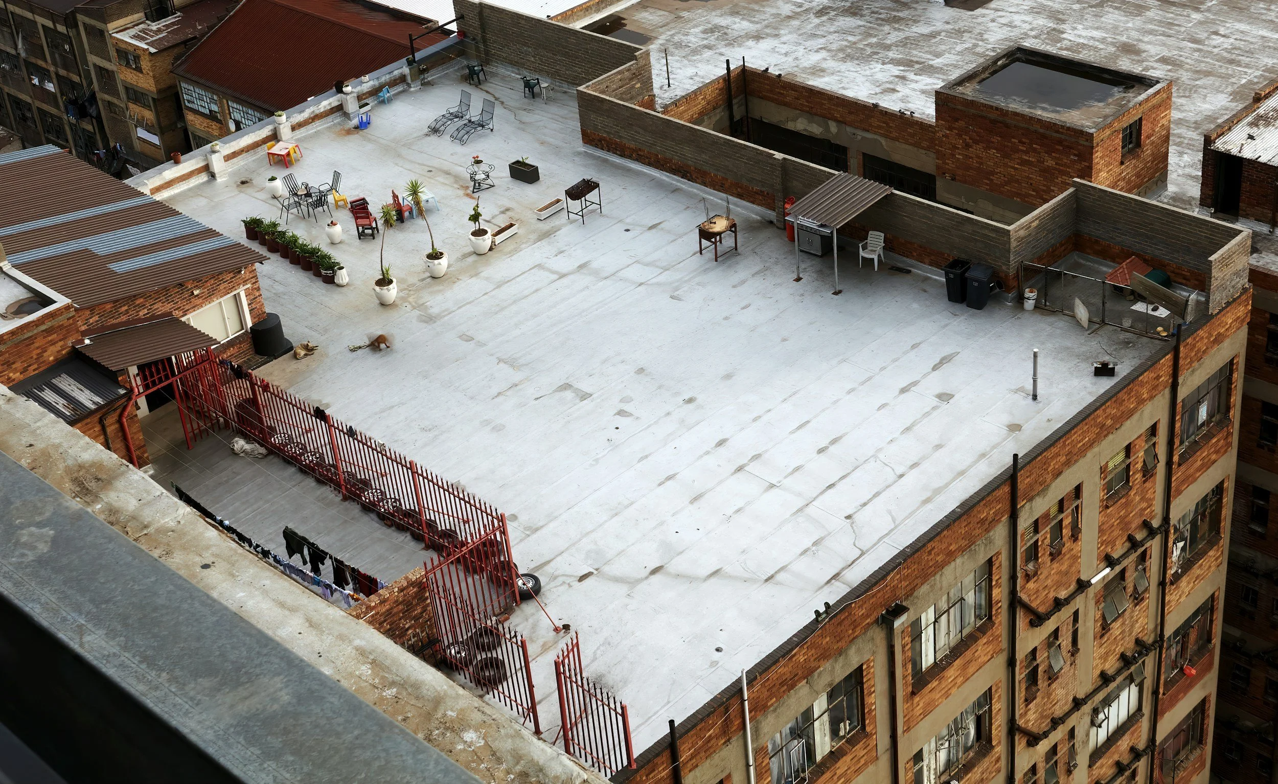 A view of a rooftop terrace in an urban area, with outdoor furniture, potted plants, a clothesline with laundry, and some scattered objects, surrounded by brick buildings.
