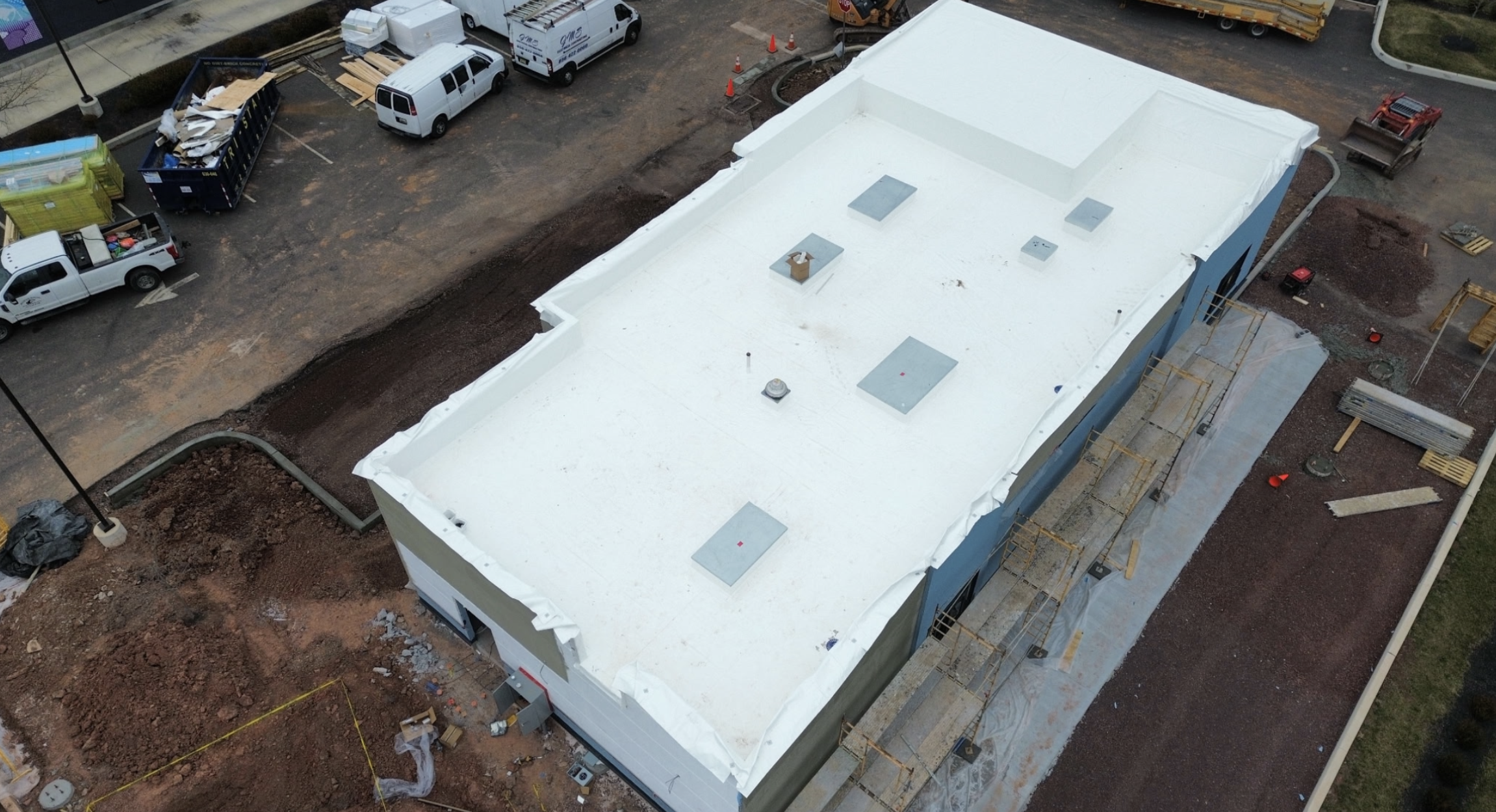 An aerial view of a construction site featuring a building with a new white roof, construction materials, equipment, and vehicles surrounding the area.
