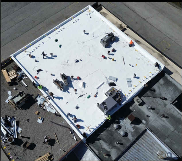 Workers installing a white roofing membrane on a large commercial building rooftop, with tools and materials scattered around.