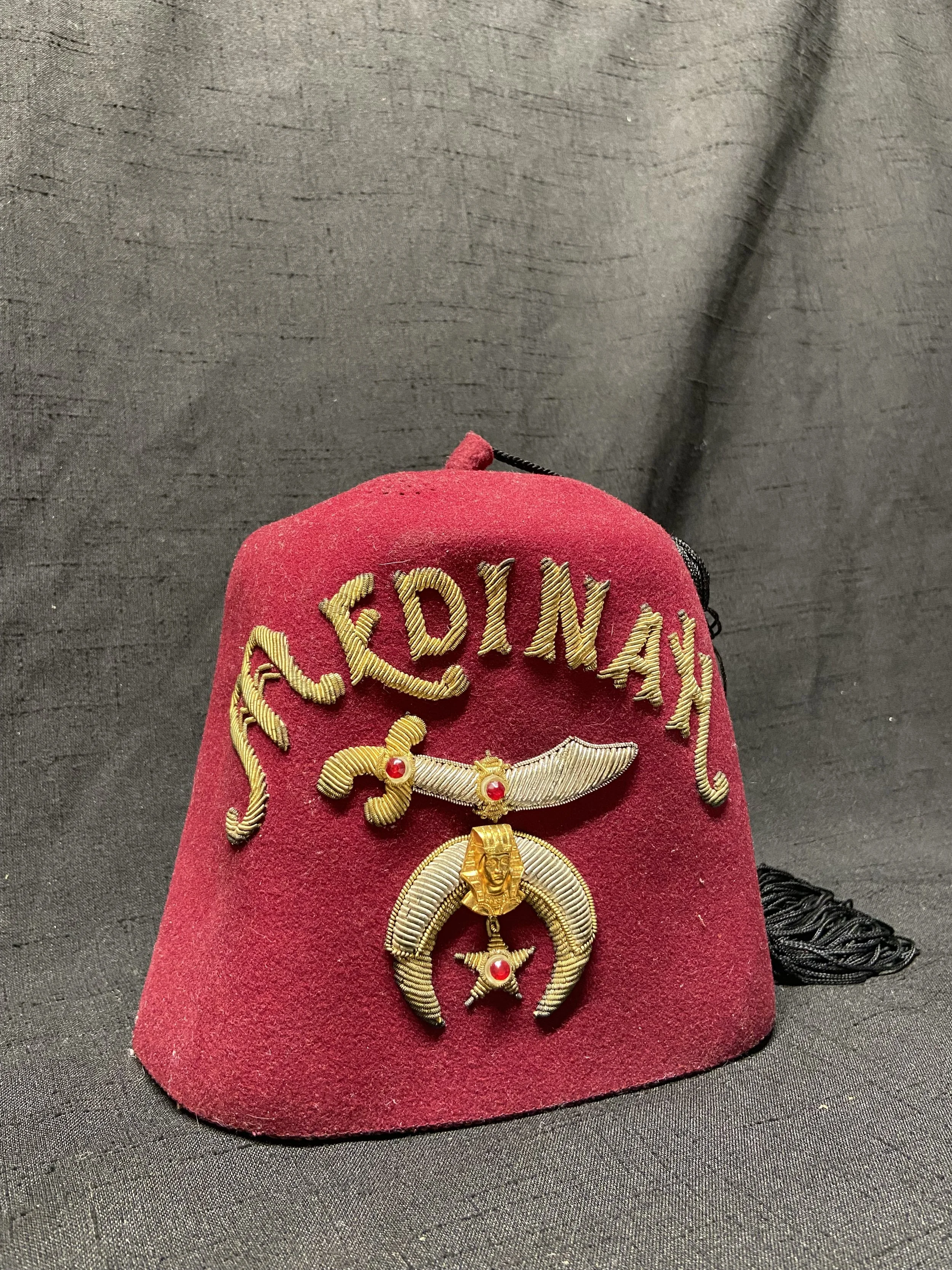 This vintage Medinah Shrine fez features the elaborate red felt construction, gold-tone bullion-style embroidery, jeweled Shrine emblem, and long black tassel associated with Medinah's ceremonial regalia.