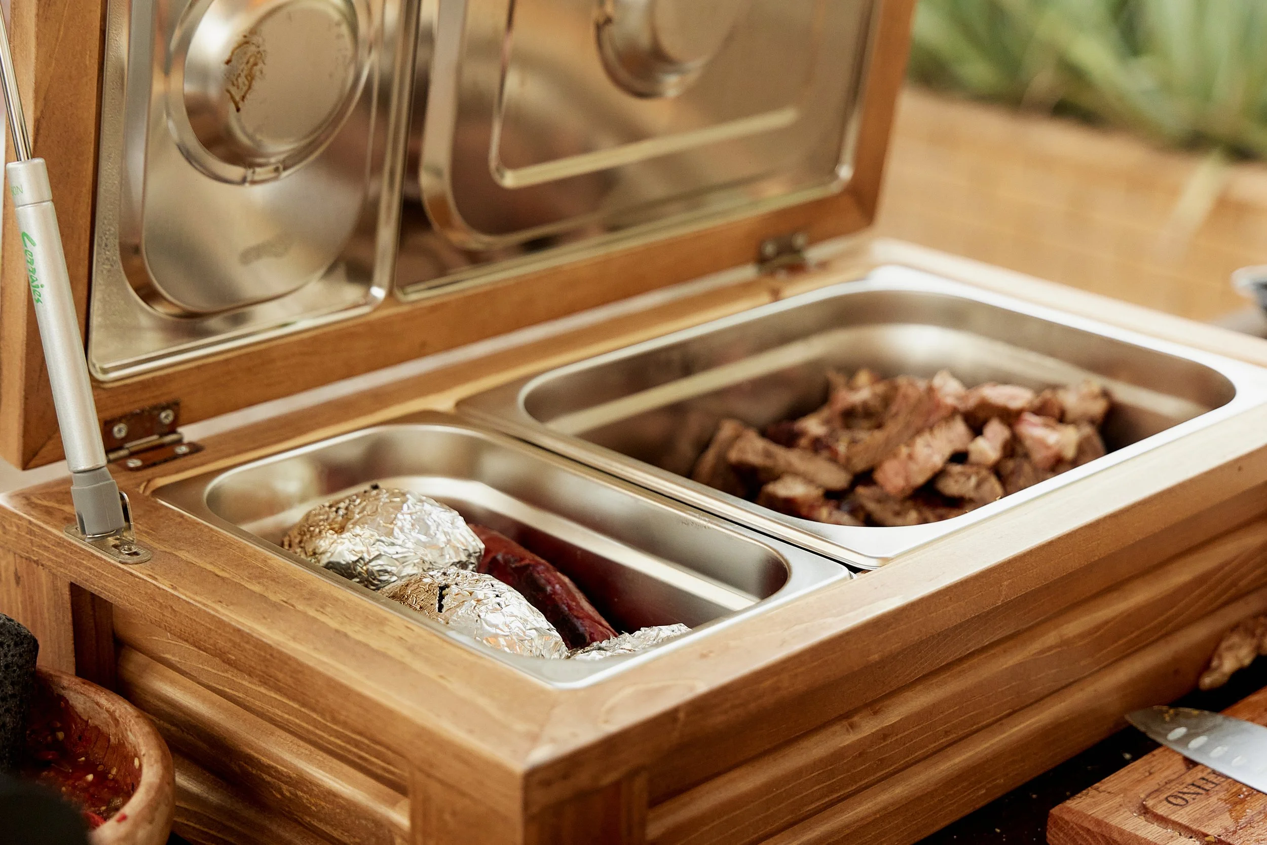 Bandeja de comida con carne y papas, en una mesa de madera con algunos recipientes de aluminio y otros de cerámica.
