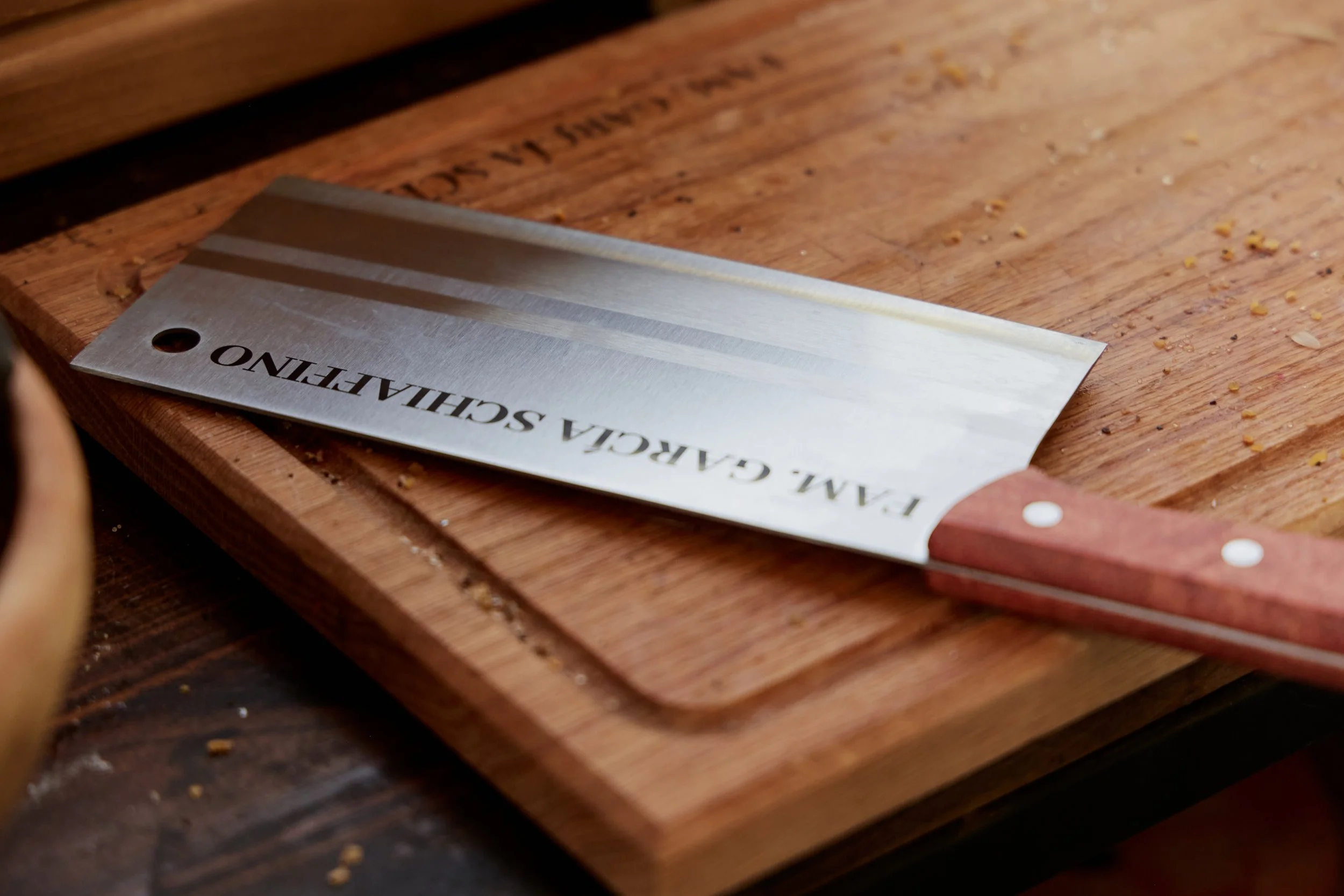 Cuchillo con hoja de acero inoxidable y mango de madera sobre una tabla de cortar de madera, con algunas migas y restos de comida.