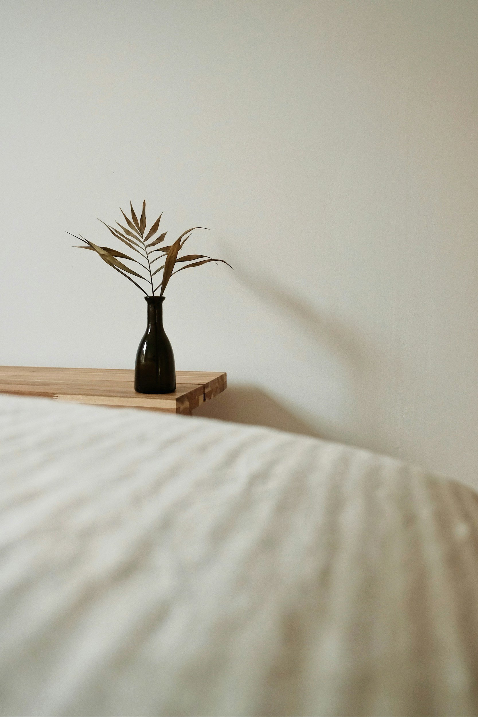 A black vase with dried palm leaves sits on a wooden surface against a plain white wall.