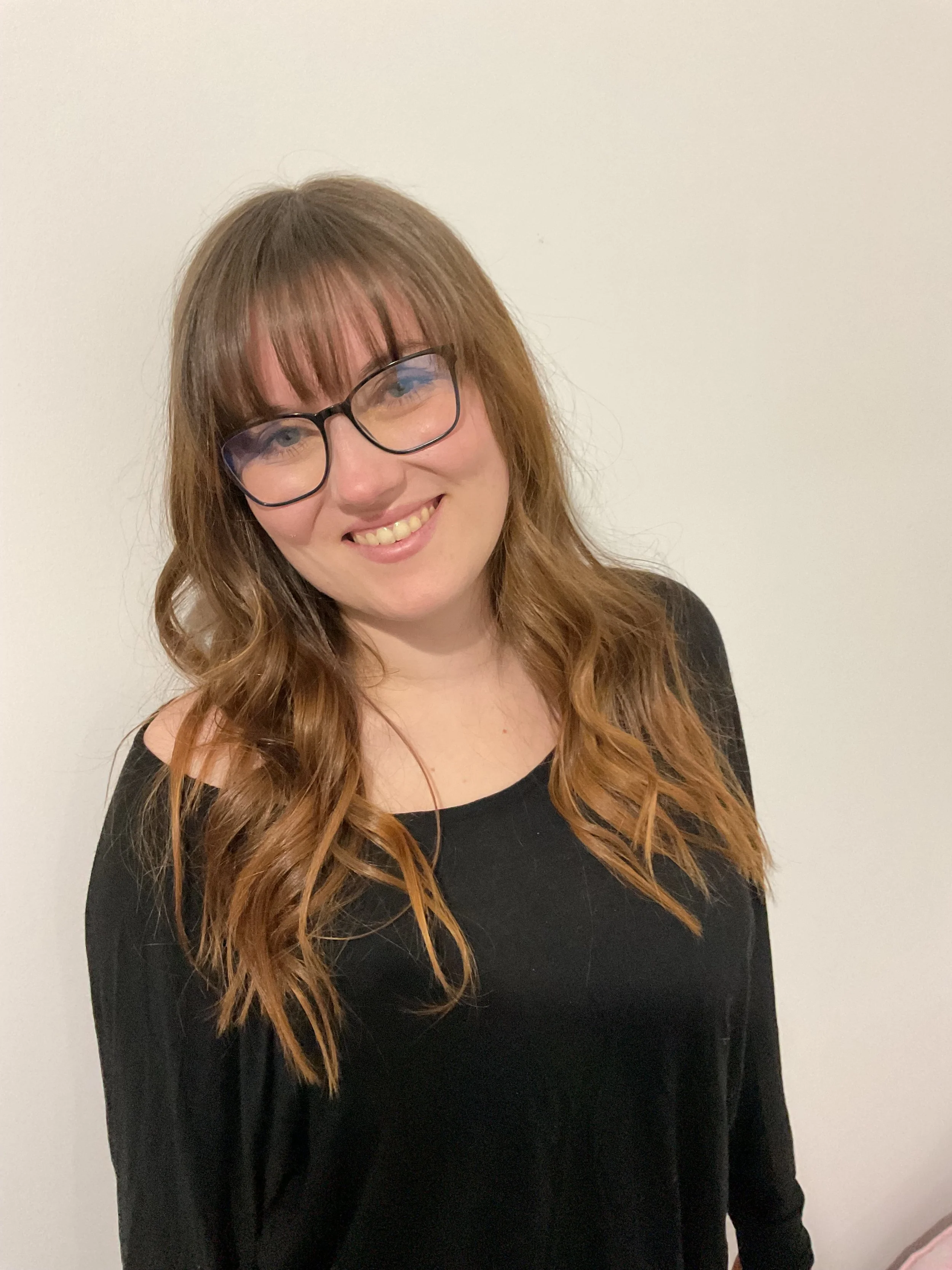 A woman with long, wavy, light brown hair wearing glasses and a black top standing in front of a plain white wall.