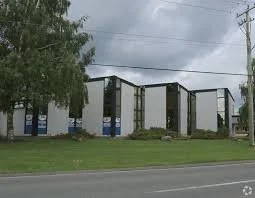 Commercial building with multiple connected sections, surrounded by grass and trees, under a cloudy sky.