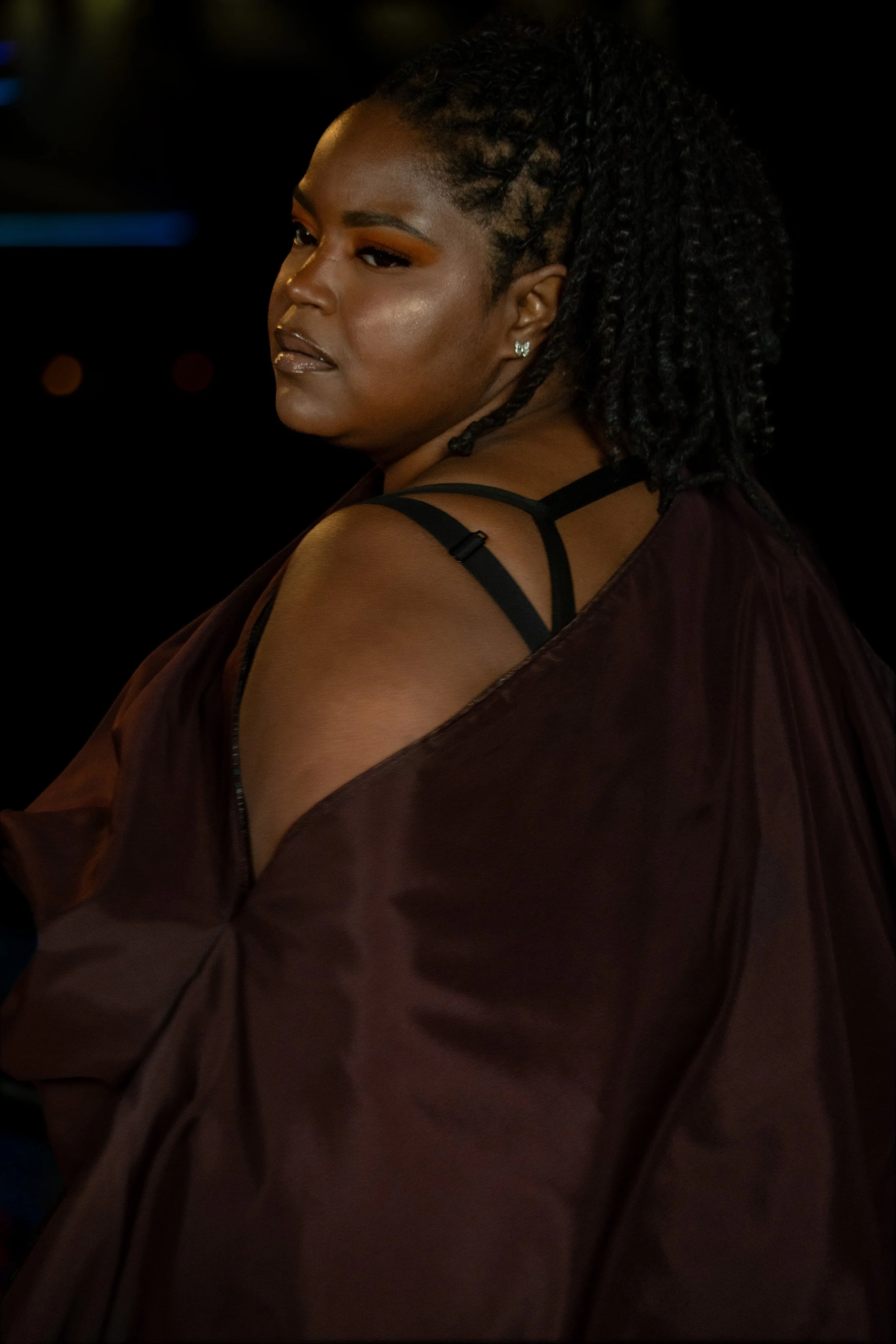 Close-up of a woman with dark skin, styled with textured braids or twists, wearing a dark-colored garment with thin straps, with a dark background.