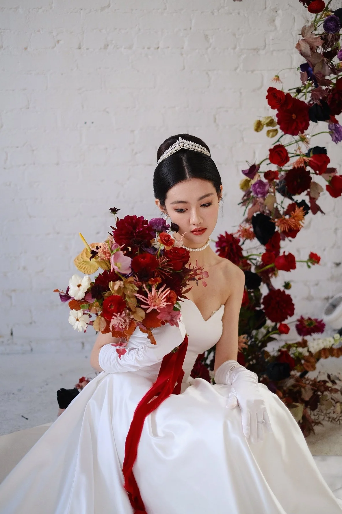 A woman in a wedding dress holding a bouquet of flowers with a white brick wall and floral arrangement in the background.
