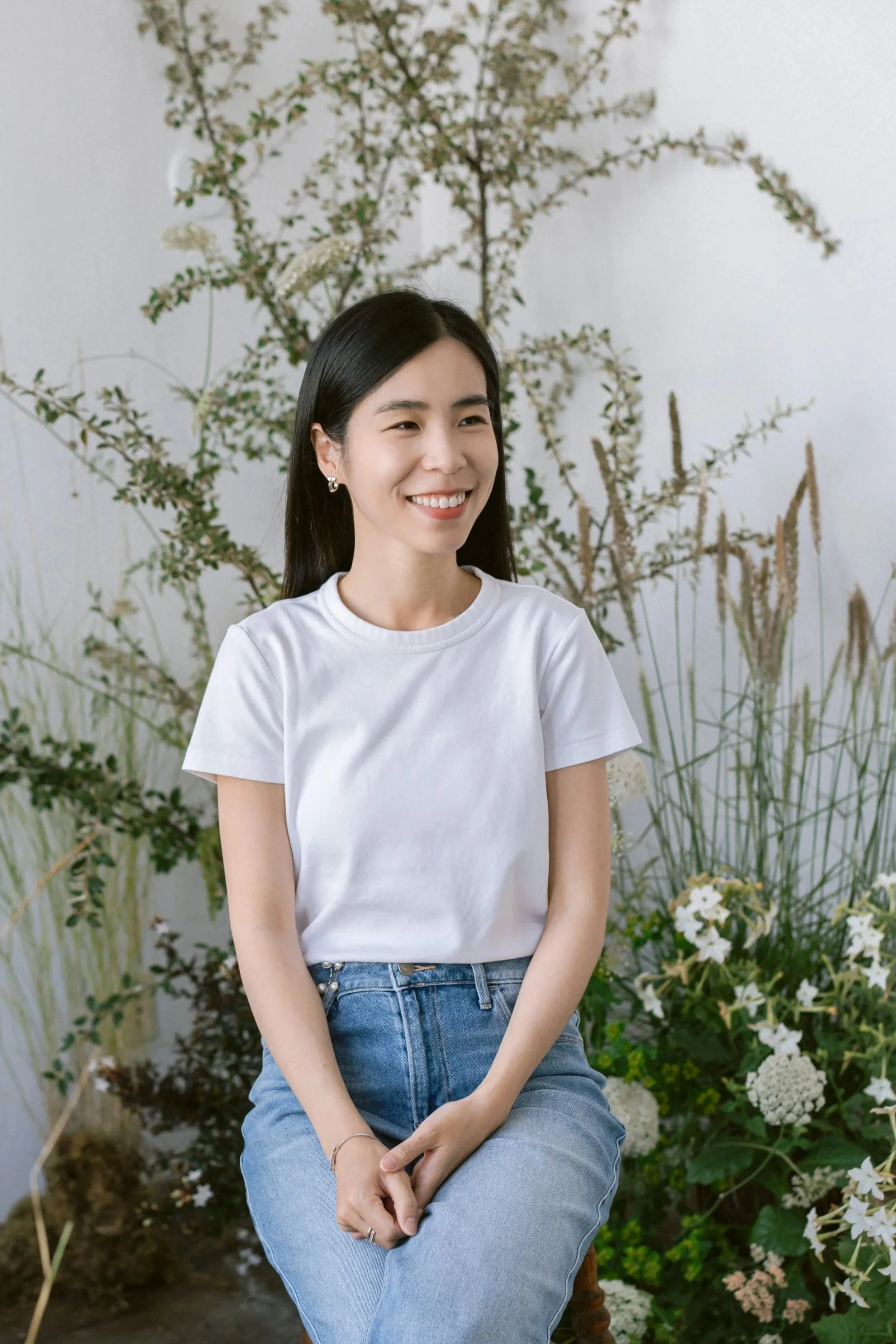 Smiling woman sitting in front of a white wall with dried and green plants, wearing a white t-shirt and blue jeans.