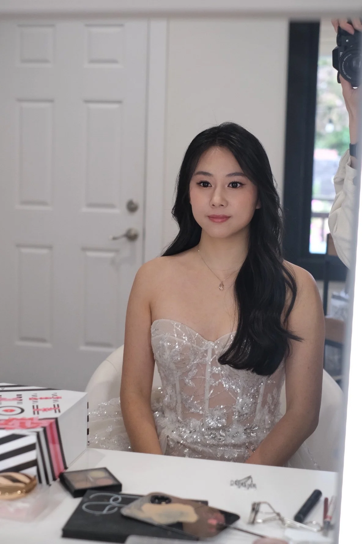 Young woman with long black hair and a strapless white dress sitting at a table with makeup products, in front of a mirror, in a room with a white door and window.