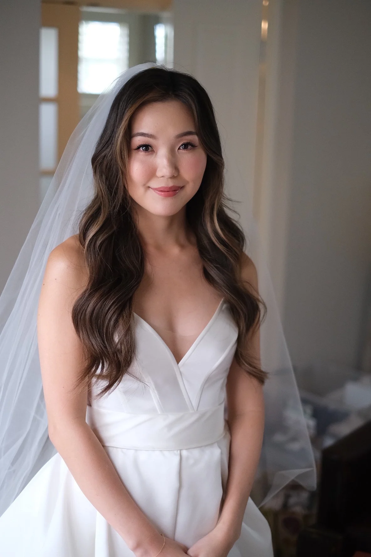 A woman in a white wedding dress with a veil standing indoors with a blurred background.