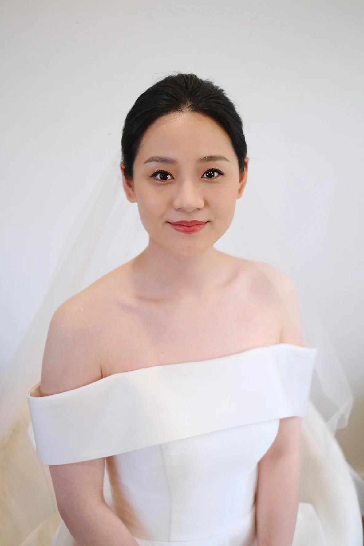 A woman with dark hair styled back, wearing a white off-the-shoulder dress and a veil, smiling with a soft expression against a plain light background.