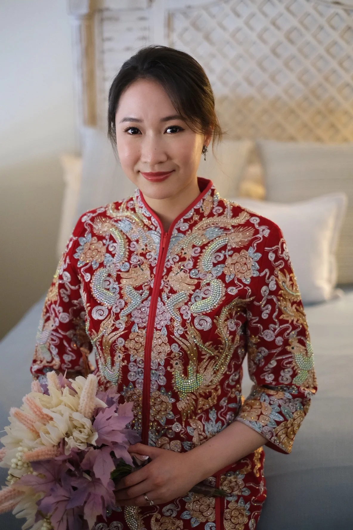 A woman in a traditional red embroidered dress holding a bouquet of pink and white flowers, standing in a bedroom with a white bed and pillows in the background.