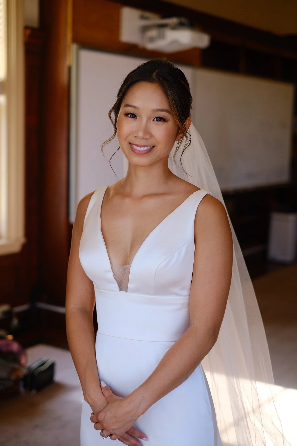 A woman dressed in a white wedding gown with a deep V neckline, standing indoors with a smile on her face and a veil on her head.