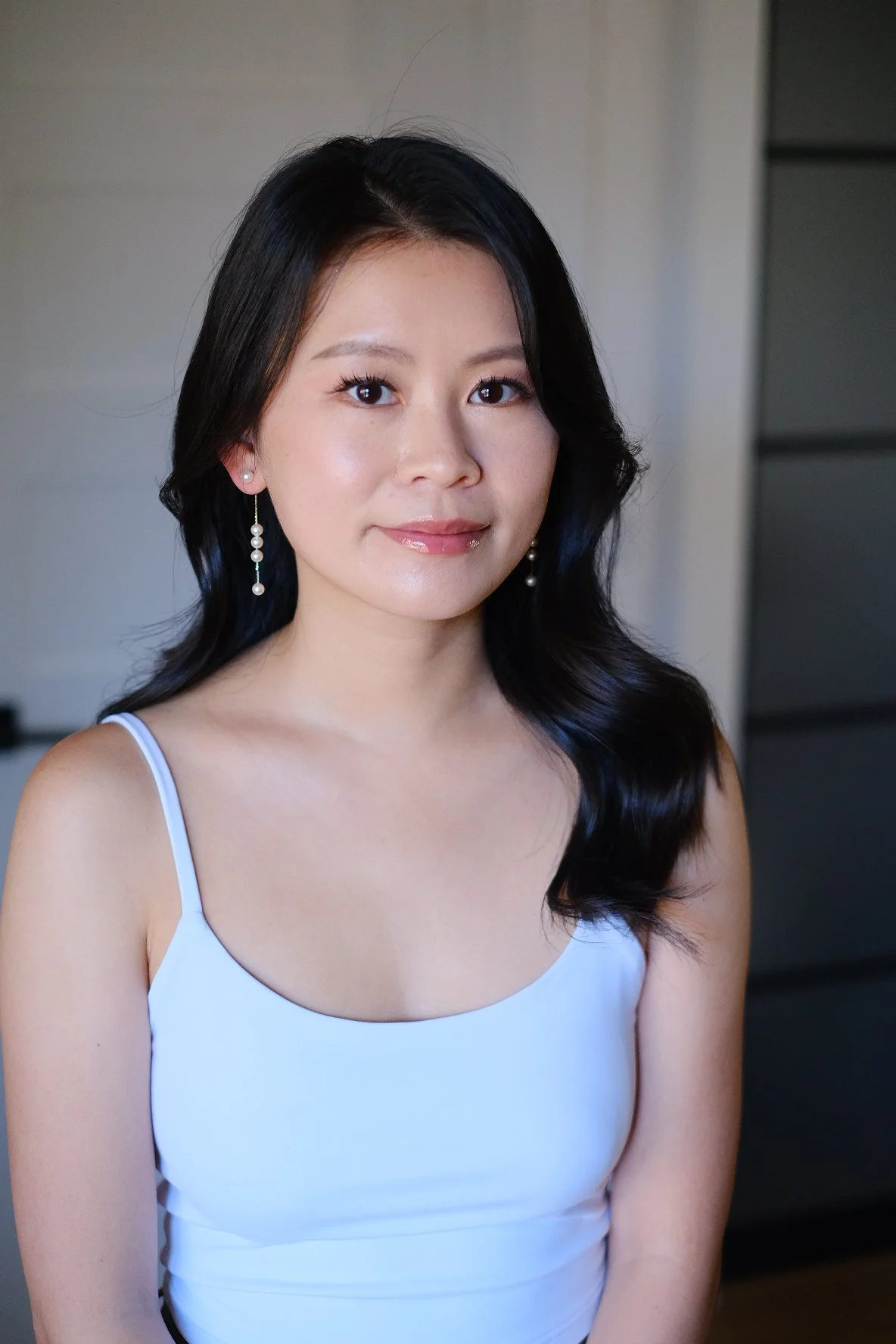 A young woman with black hair styled in soft waves, wearing pearl earrings and a white dress with thin straps, standing indoors against a neutral background.