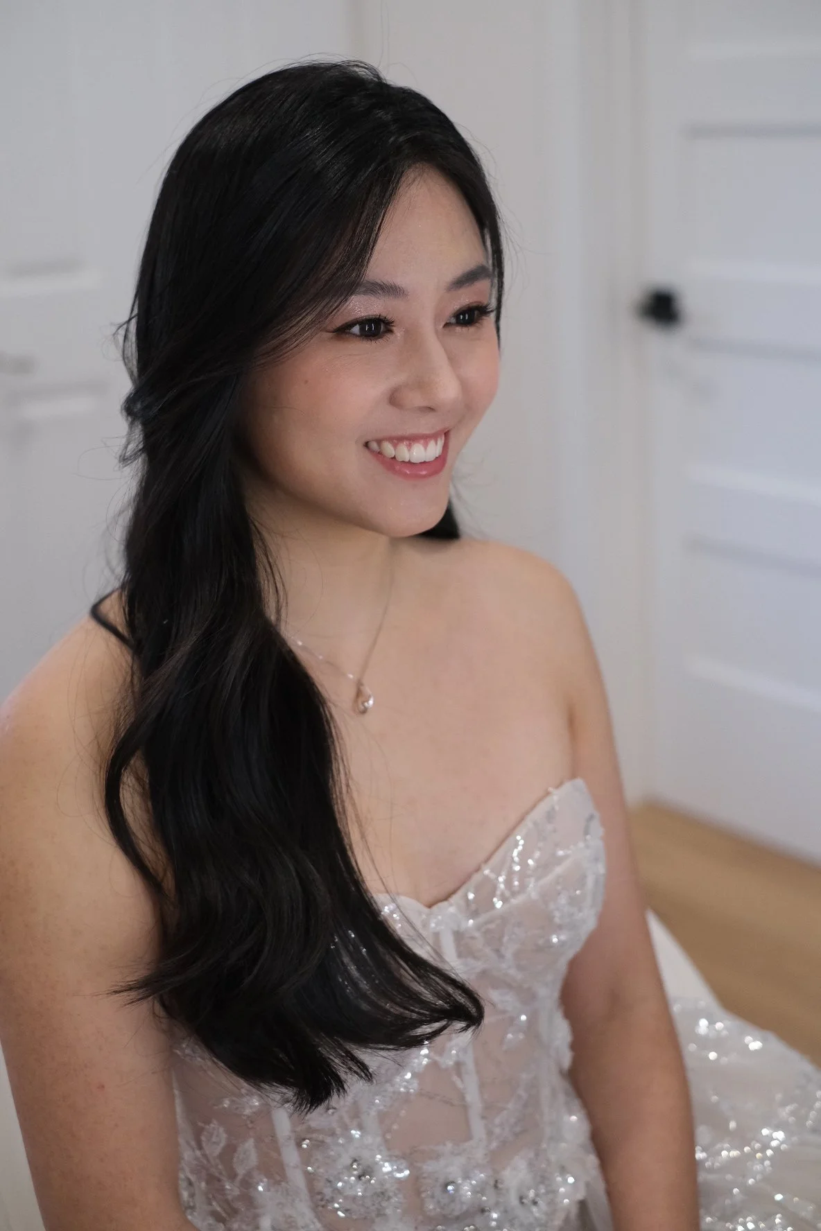 A woman with long black hair in loose curls wearing a silver wedding dress with lace and sequin details, a necklace, and smiling with visible teeth, sitting indoors with white walls and furniture in the background.