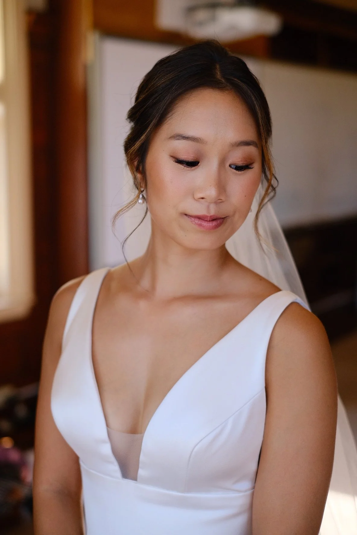 A woman in a white wedding dress with a deep V-neckline, looking down with a gentle smile, in an indoor setting.