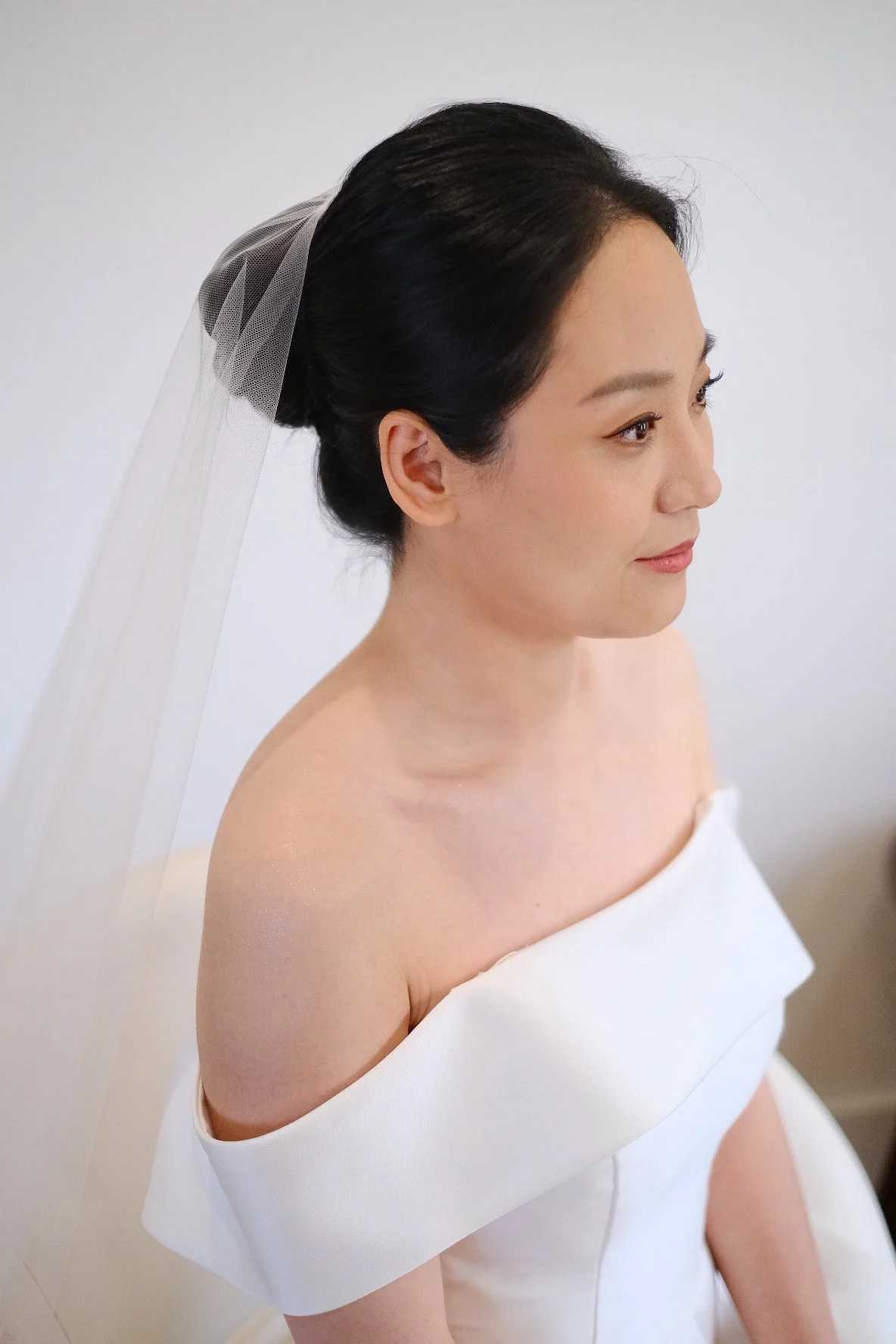 A woman with dark hair styled in an elegant updo wearing a white off-shoulder dress and a white veil against a plain light background.