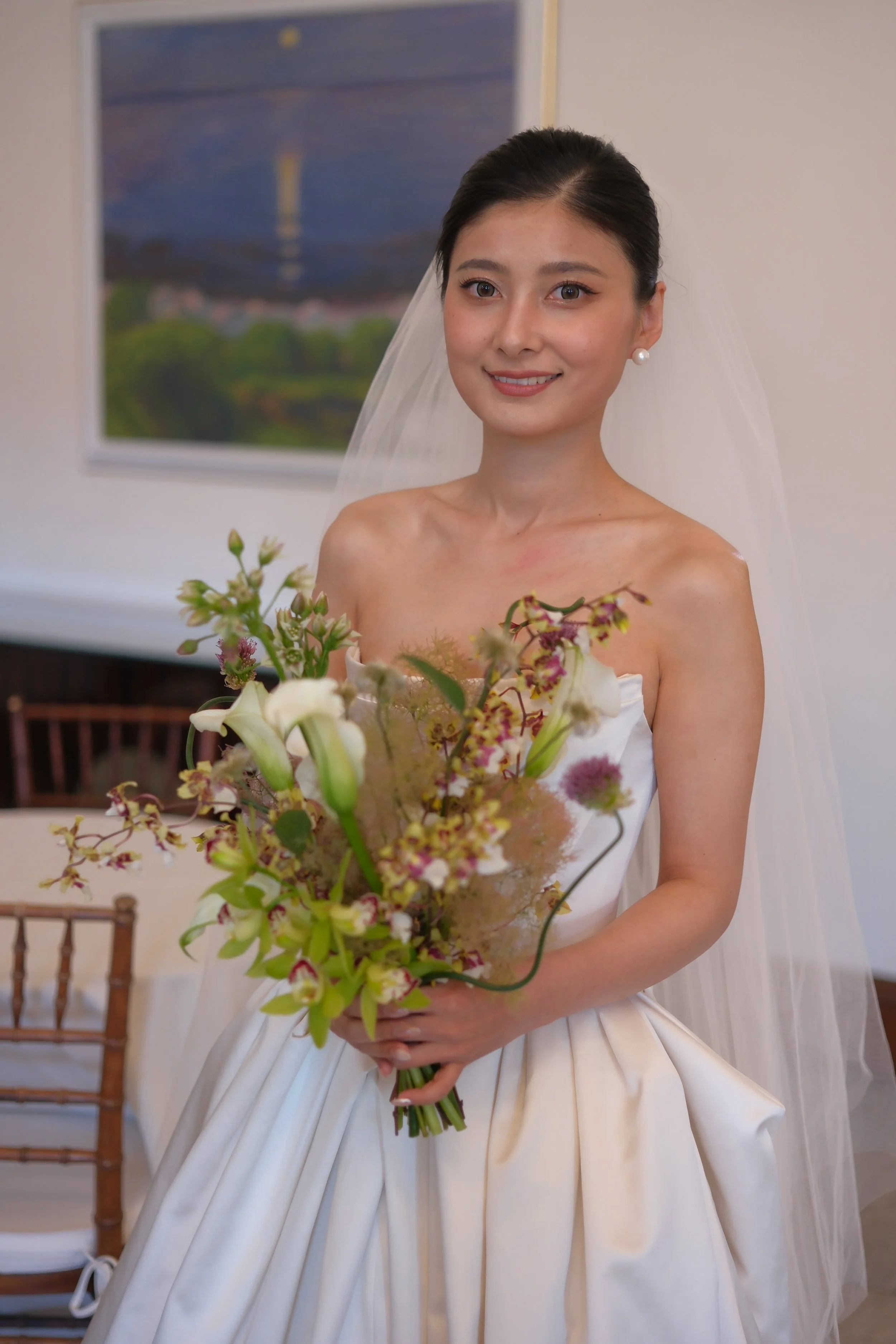 A smiling woman dressed as a bride holding a bouquet of flowers, wearing a white wedding gown and veil, with a framed landscape painting on the wall in the background.