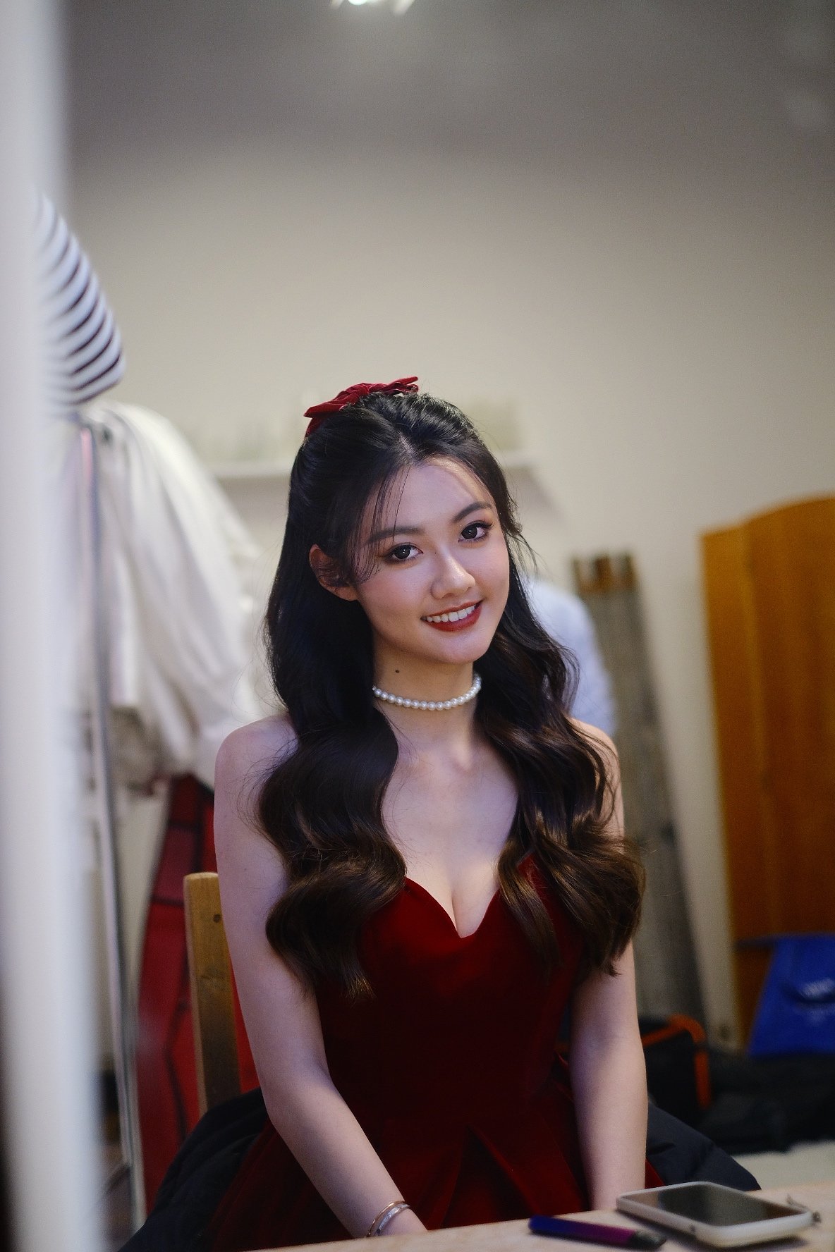A young woman with long wavy dark hair, wearing a red dress, pearl necklace, and makeup, smiling while sitting in an indoor setting with a blurred background.