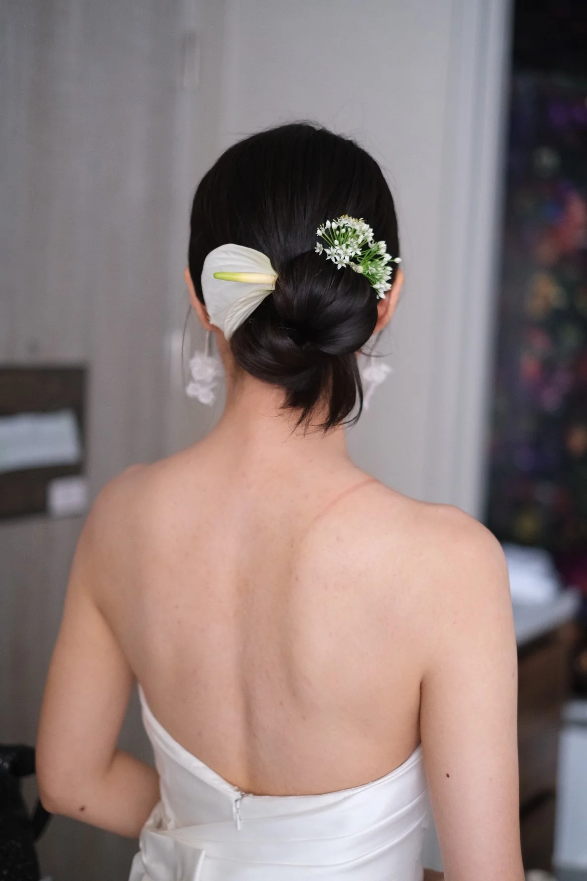 Back view of a woman with dark hair styled in an elegant bun, adorned with white flowers and a white leaf, wearing a strapless white dress.