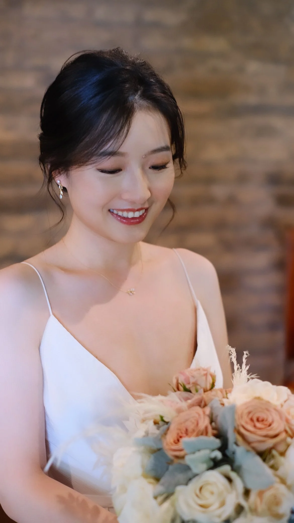 A woman wearing a white dress and jewelry, holding a bouquet of pink and white flowers, smiling and looking down.