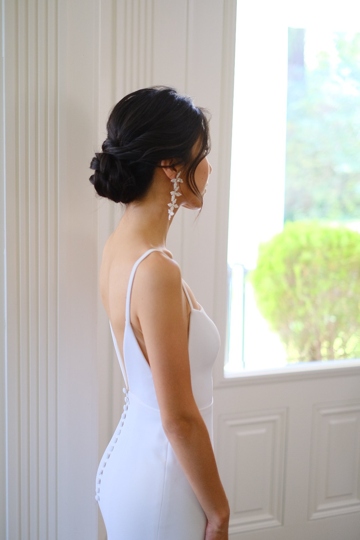 A woman with dark hair styled in an elegant updo, wearing a white dress and long floral earrings, standing near a window with greenery outside.