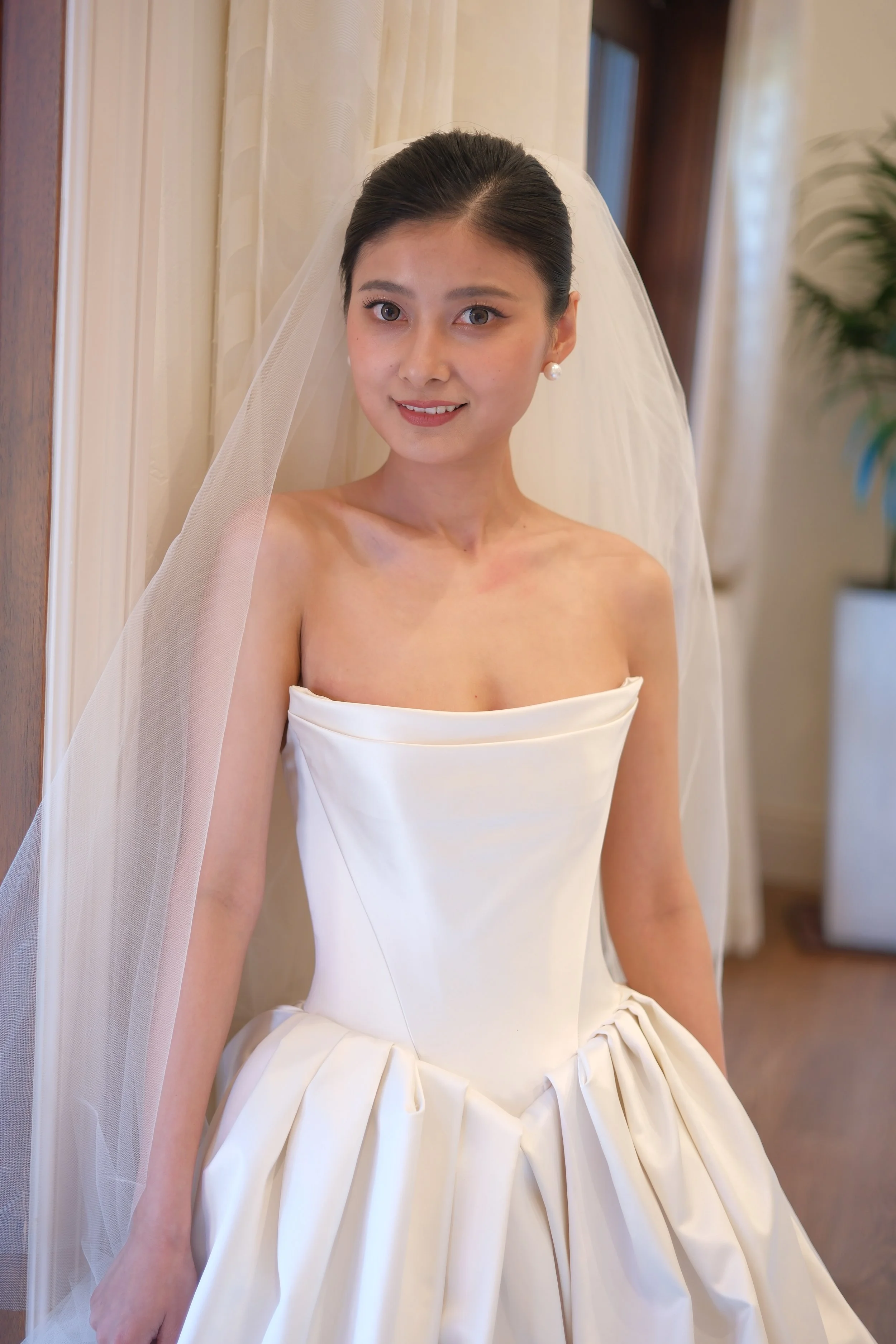 A woman dressed in a strapless white wedding gown with a full skirt, posed indoors near a doorway with curtains. She has a veil and pearl earrings, and is smiling softly.