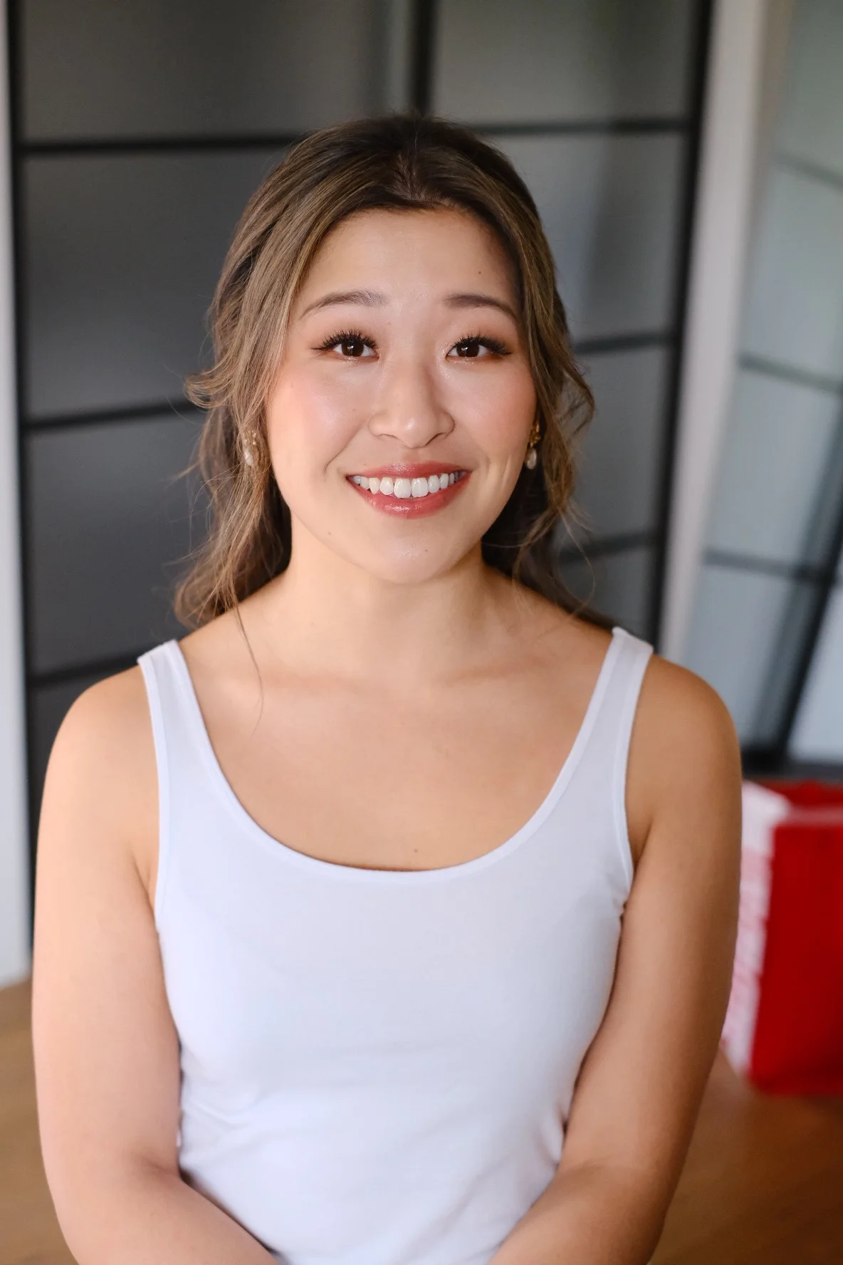A young woman with shoulder-length wavy hair, smiling, wearing a white tank top, and posing indoors with a dark wall and red object in the background.