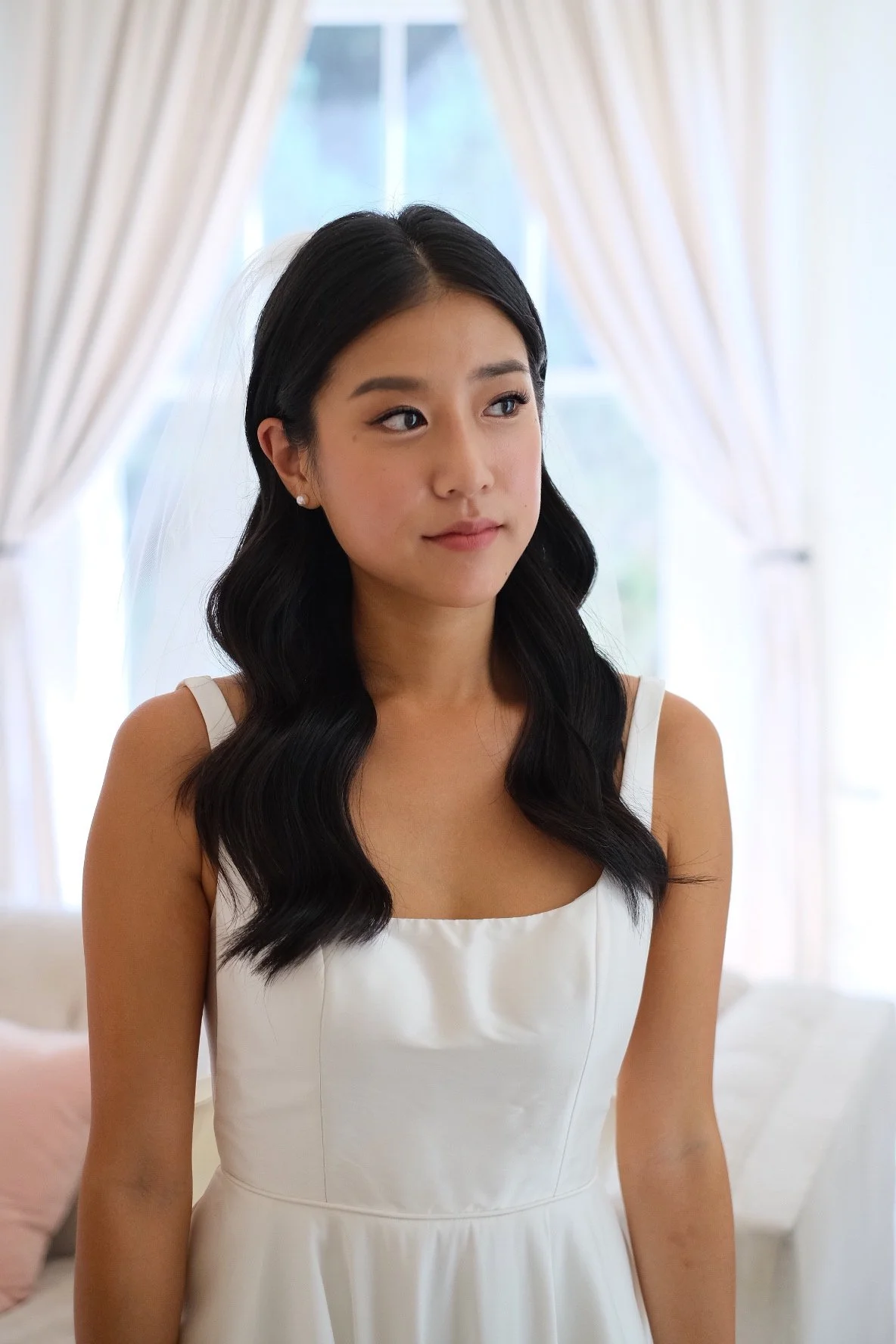 Young woman in a white dress with dark wavy hair, standing indoors near a window with beige curtains.