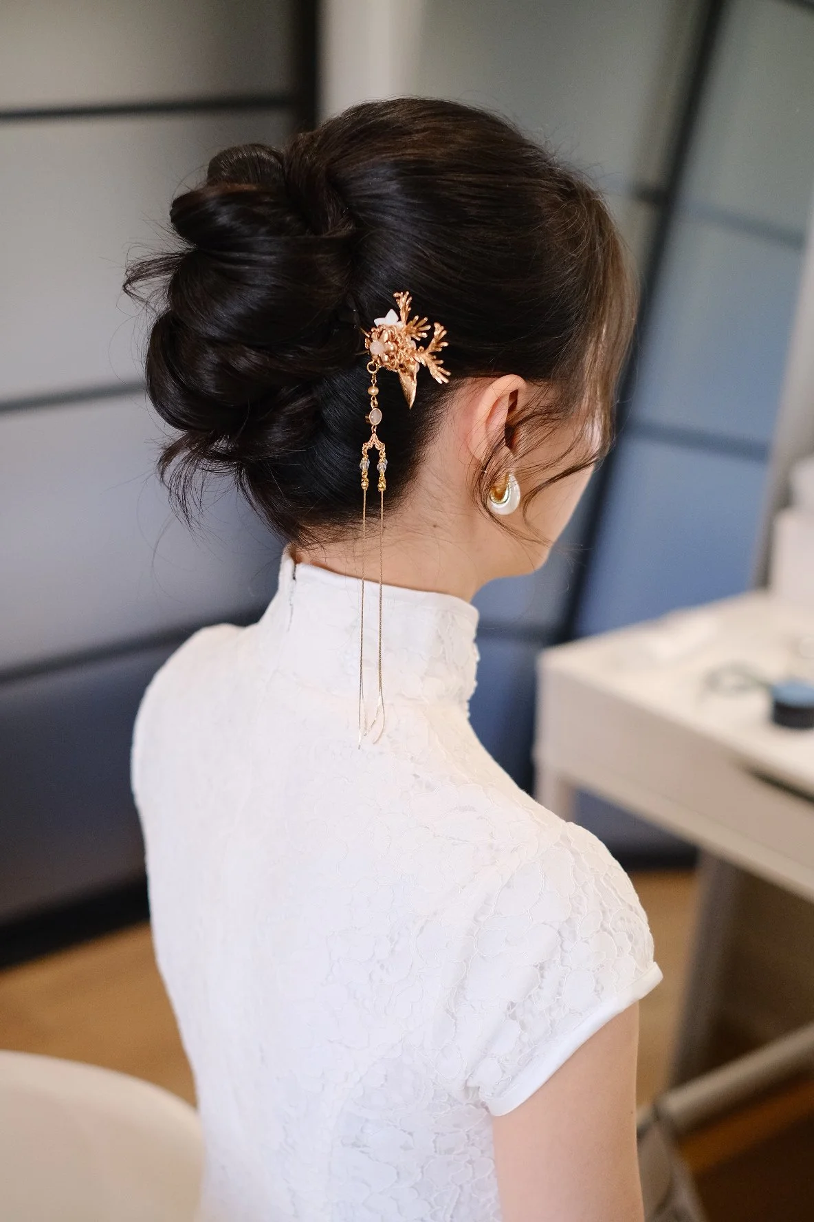 Side view of a woman with dark hair styled in an elegant updo, wearing a white textured dress, a pearl earring, and a decorative gold hair accessory with dangling chains.