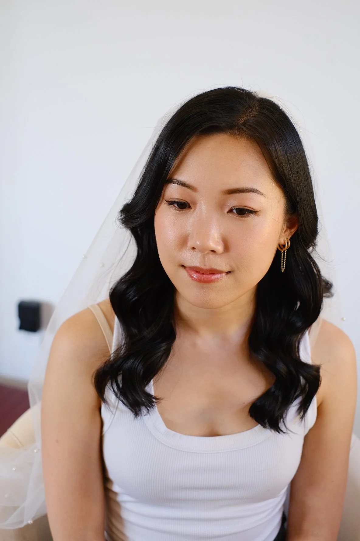 A young woman with black hair styled in loose curls, wearing gold hoop earrings, and a white sleeveless top, looking down slightly.