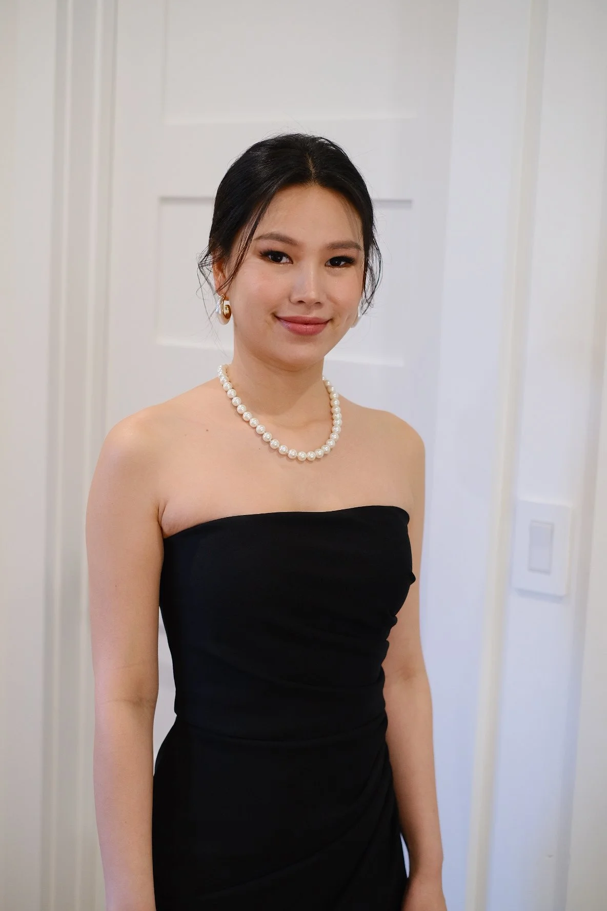A woman in a strapless black dress with pearl jewelry, smiling in front of a plain white door.