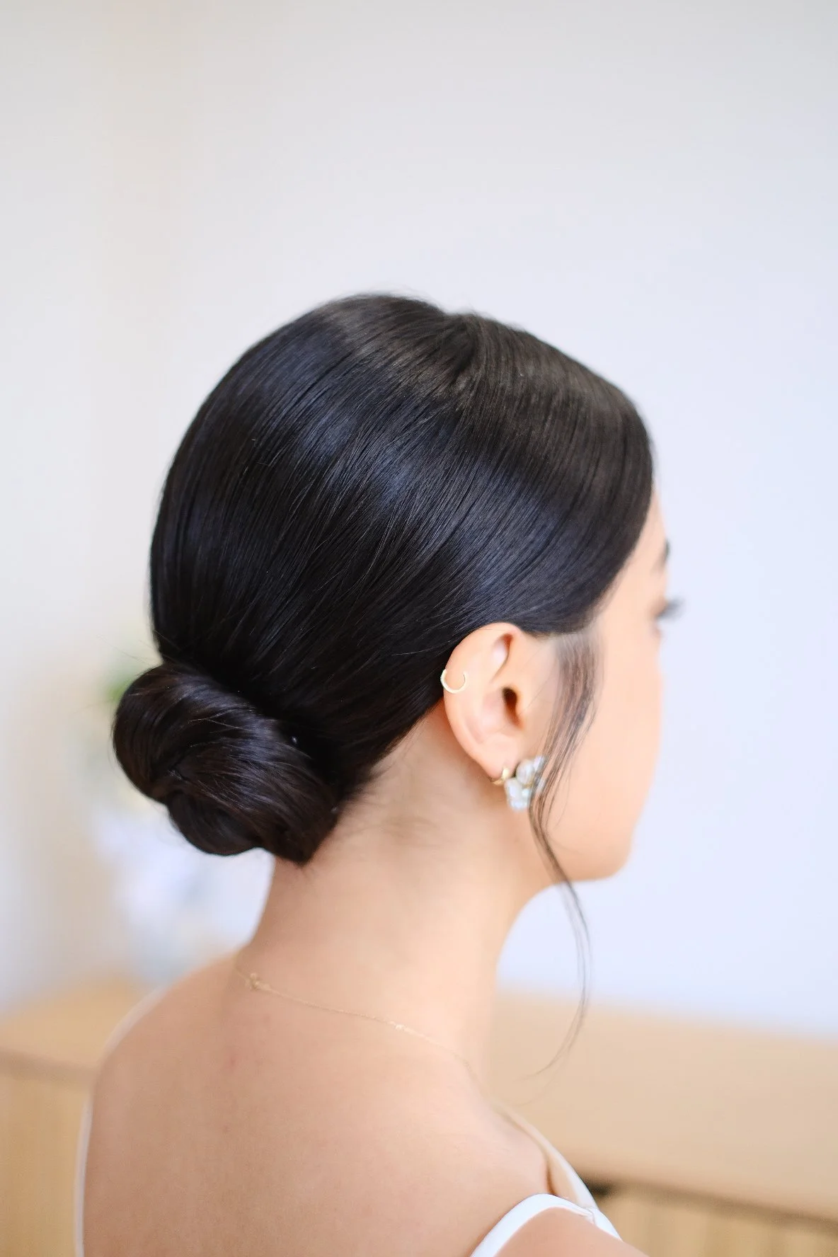 Side profile of a woman with sleek, black hair styled into a low, elegant bun, wearing small hoop earrings with white gemstone accents.