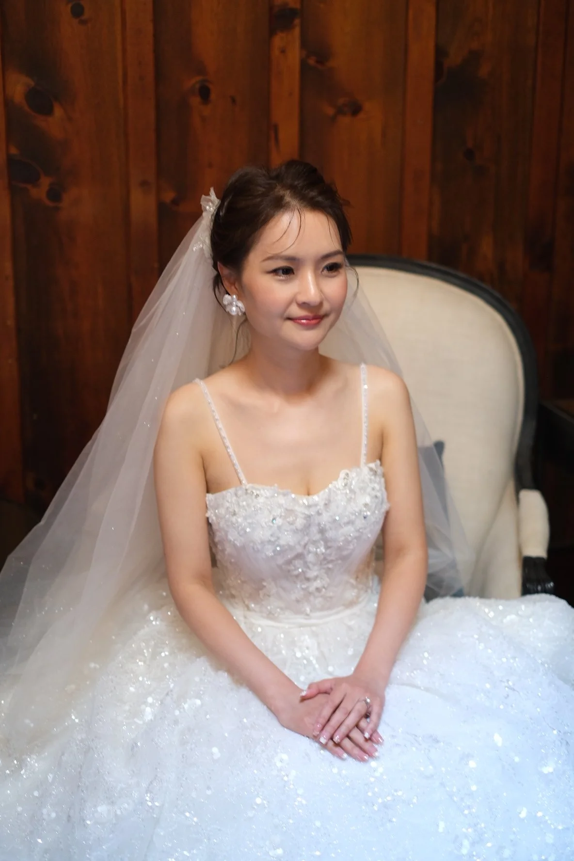 A bride sitting on a white chair in a wedding dress with a veil, smiling softly, with a wooden wall in the background.